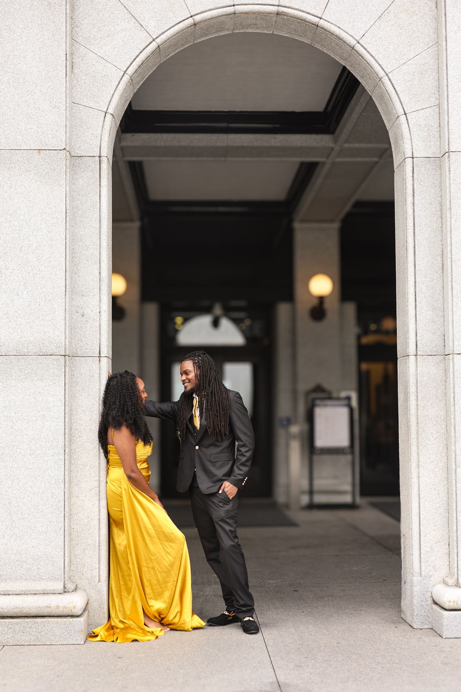 A couple shares a romantic moment under an elegant stone archway, with her in a flowing yellow gown and him in a dark suit.