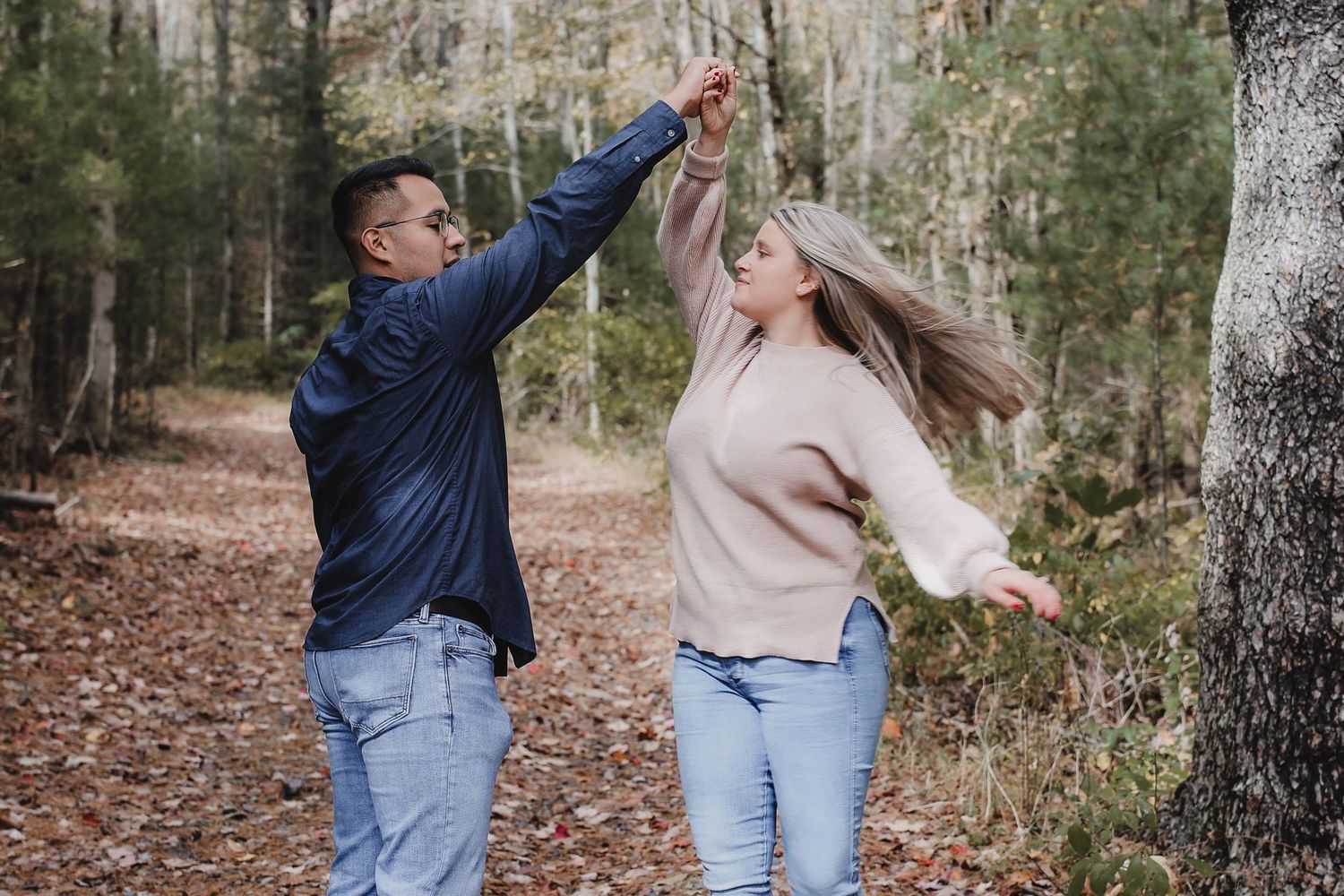 Couple in casual denim and sweaters dancing playfully through autumn forest with fallen leaves.