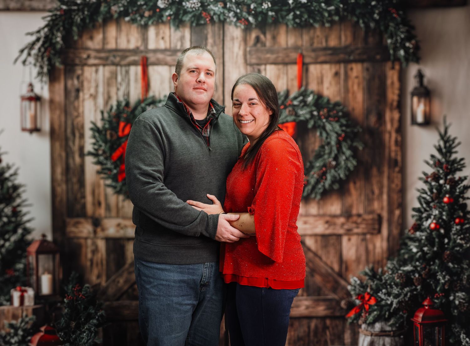 A couple in grey and red sweaters pose for holiday photos in front of a rustic wooden door with Christmas decor.