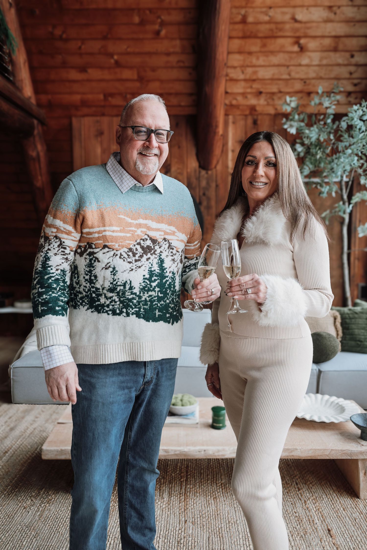 Two people in winter sweaters holding wine glasses stand together in a cozy wooden cabin interior.