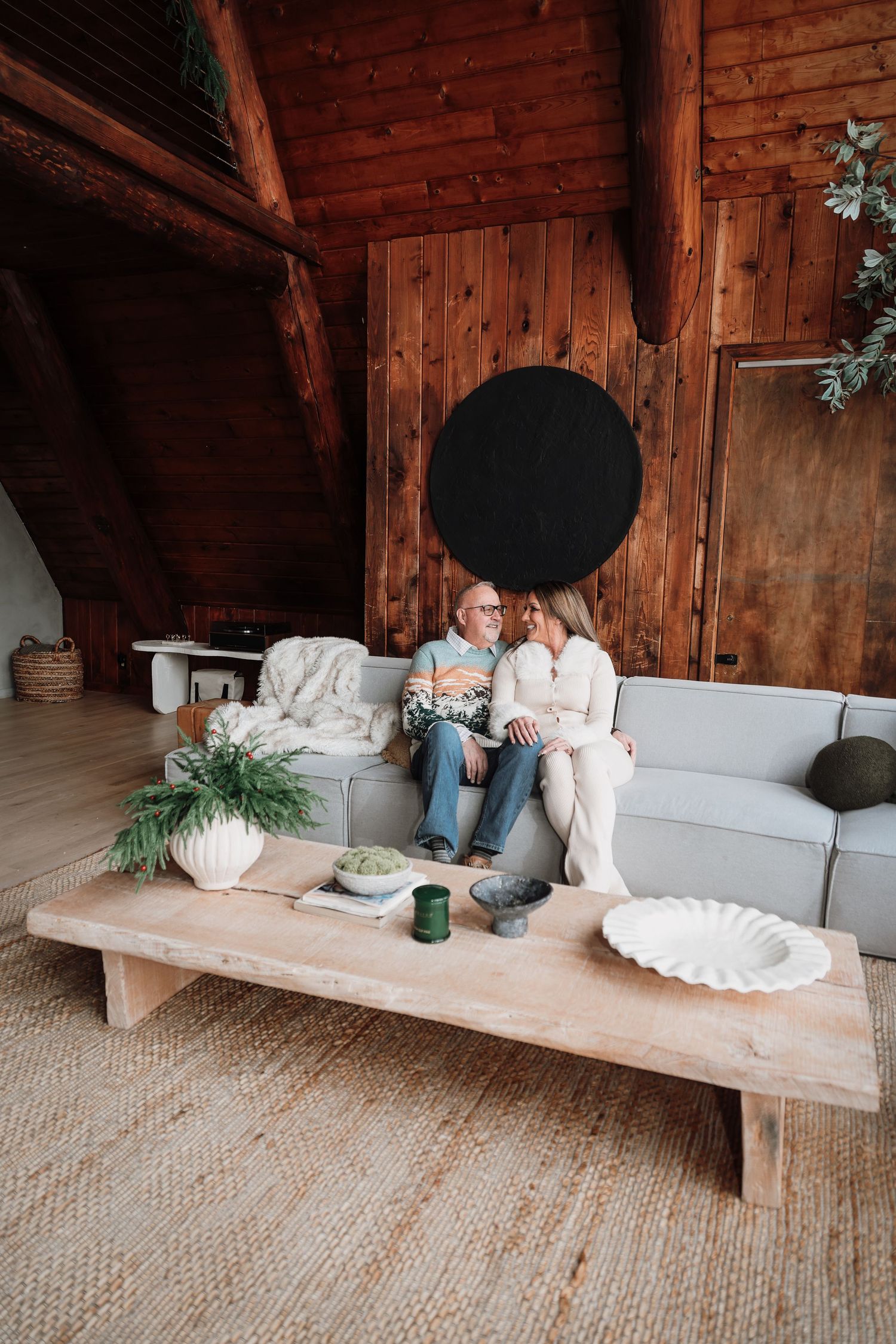 A couple relaxes together on a rustic wooden bench inside a cozy cabin with exposed wood beams and gray cushions.