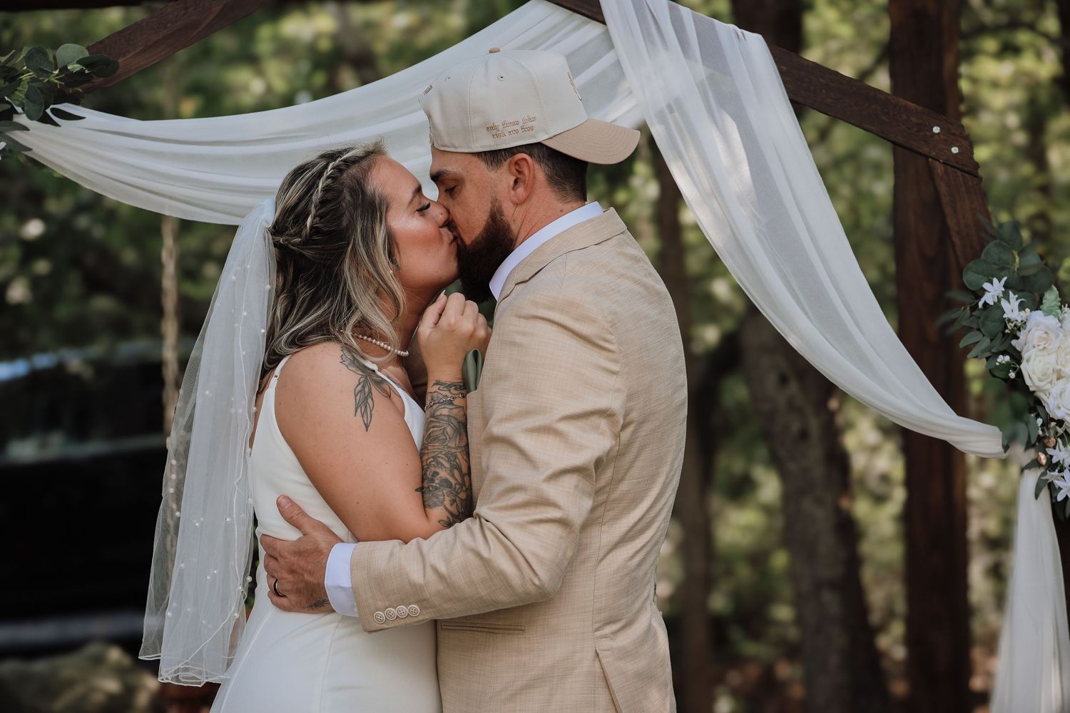 A couple shares a romantic kiss under flowing white fabric draped between trees during an outdoor wedding ceremony.