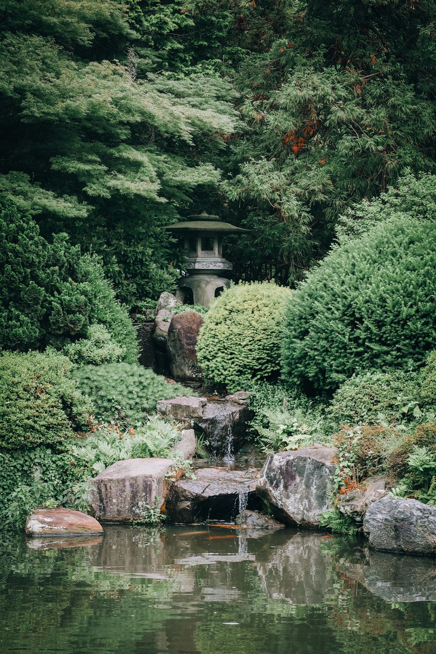 Serene Japanese garden with stone lantern, lush greenery, cascading waterfall, and tranquil pond reflecting surrounding trees.