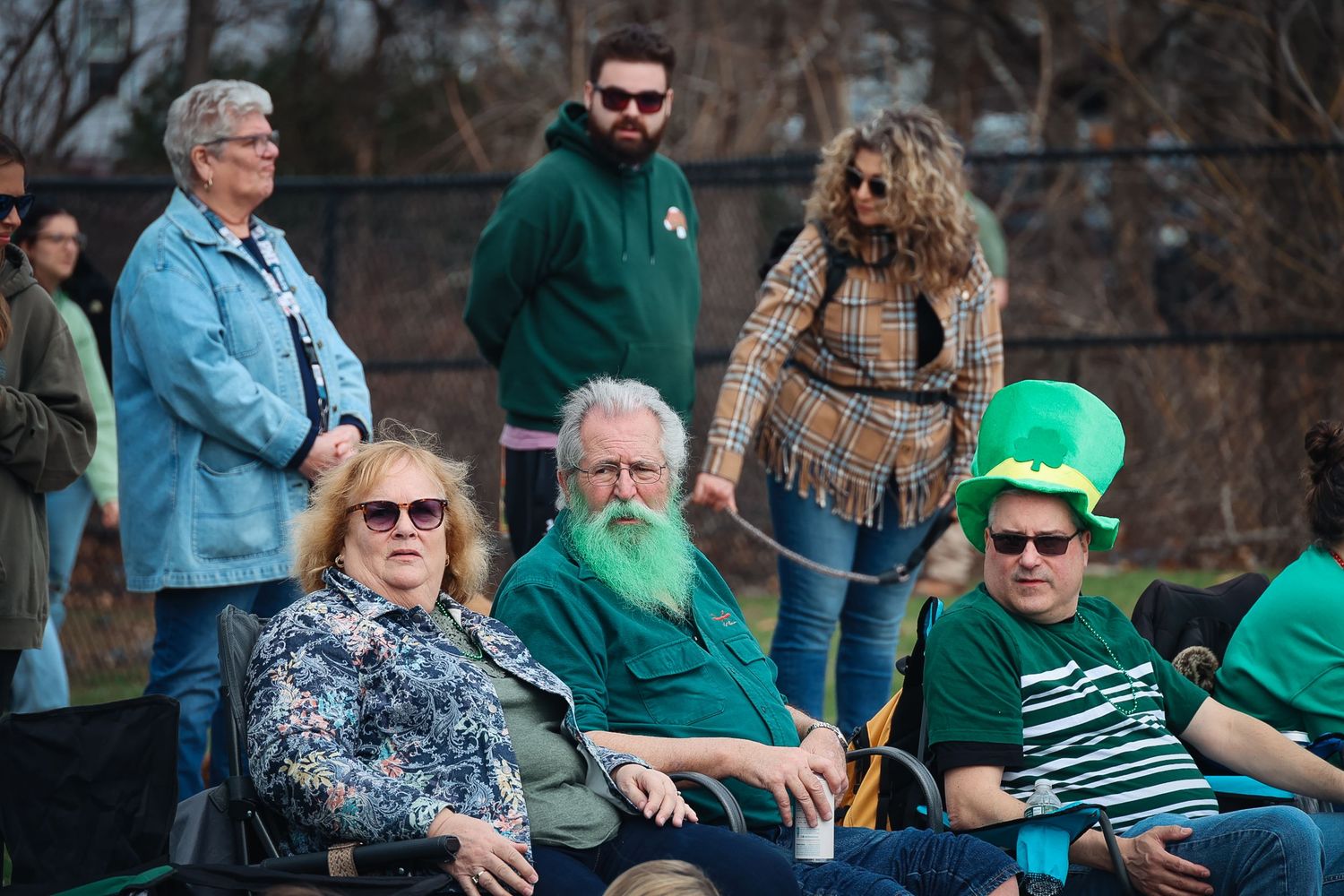 Group of people at a St. Patrick's Day parade, one man wearing a green leprechaun hat and another with a dyed green beard.