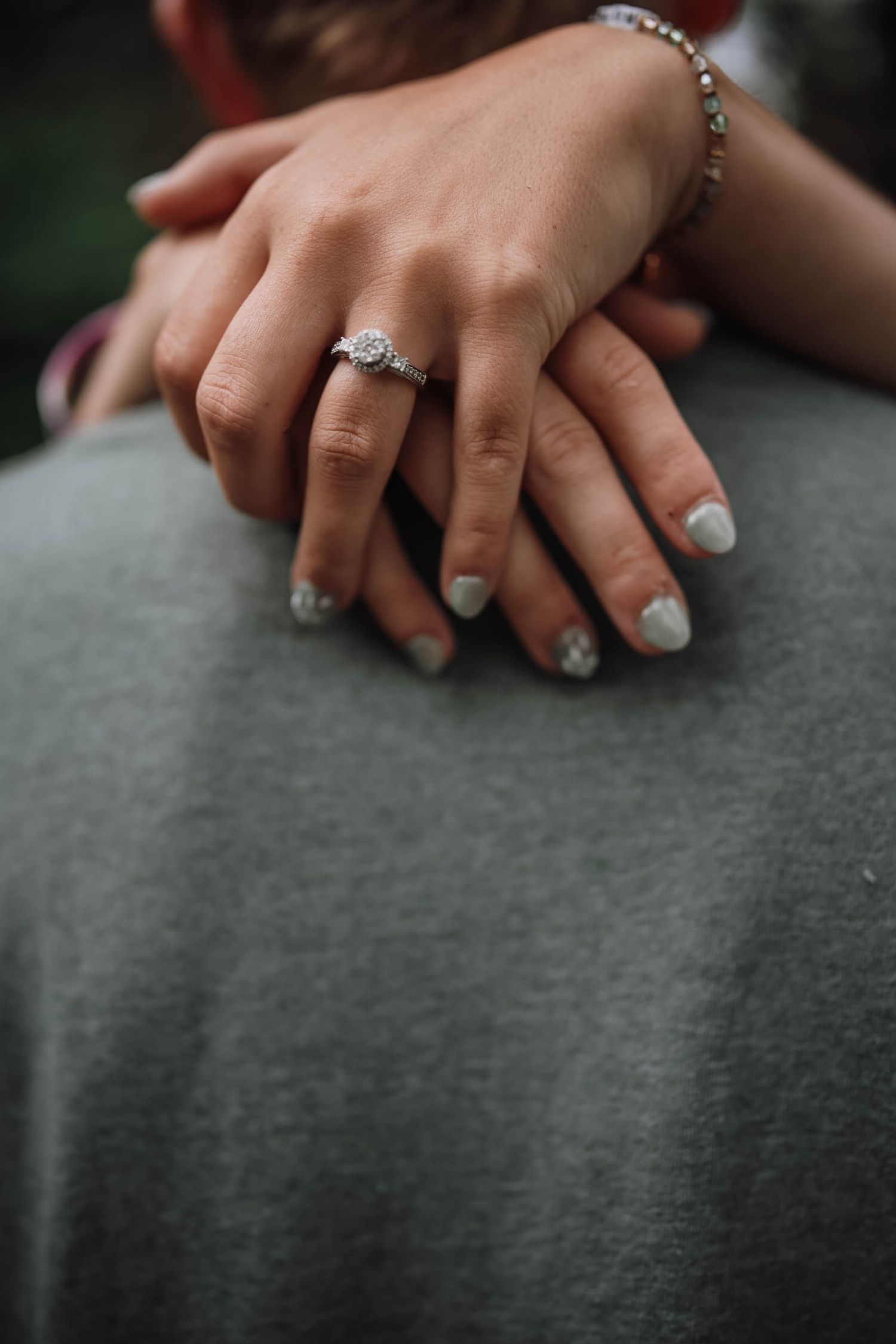 Close-up of a woman's hand with an engagement ring and bracelet resting on a man's gray shirt.