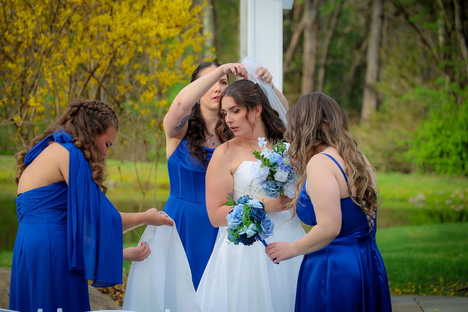 Bride in white gown holds blue bouquet as bridesmaids in royal blue dresses adjust her veil outdoors.