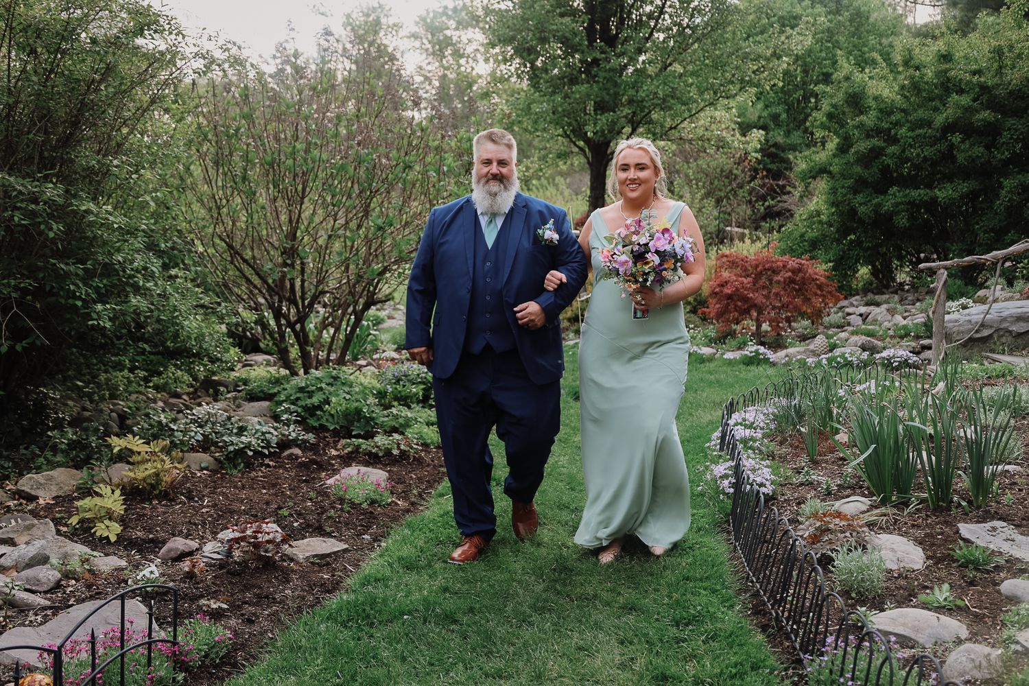 Bride in sage green gown walks with bearded groomsman in navy suit through lush garden wedding venue.