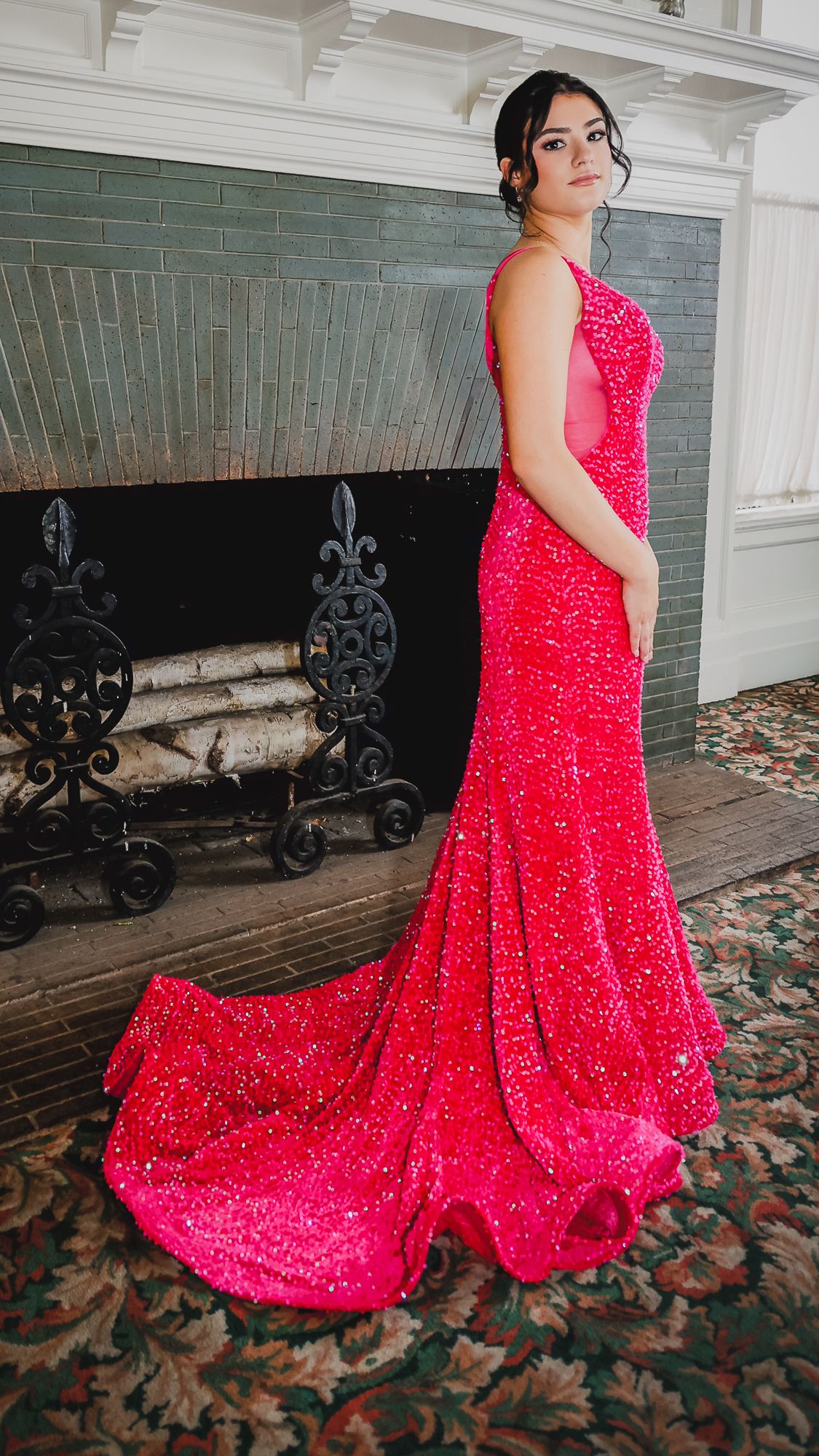 Woman in a stunning hot pink sequined mermaid gown with a train, posing elegantly in front of a fireplace.
