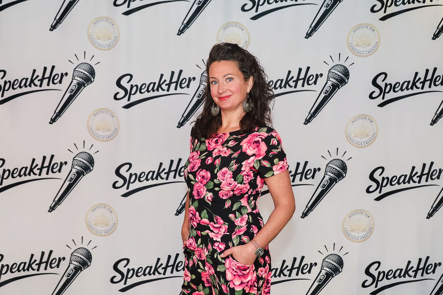 Woman in floral dress posing at SpeakHer event backdrop with microphone logo branding.