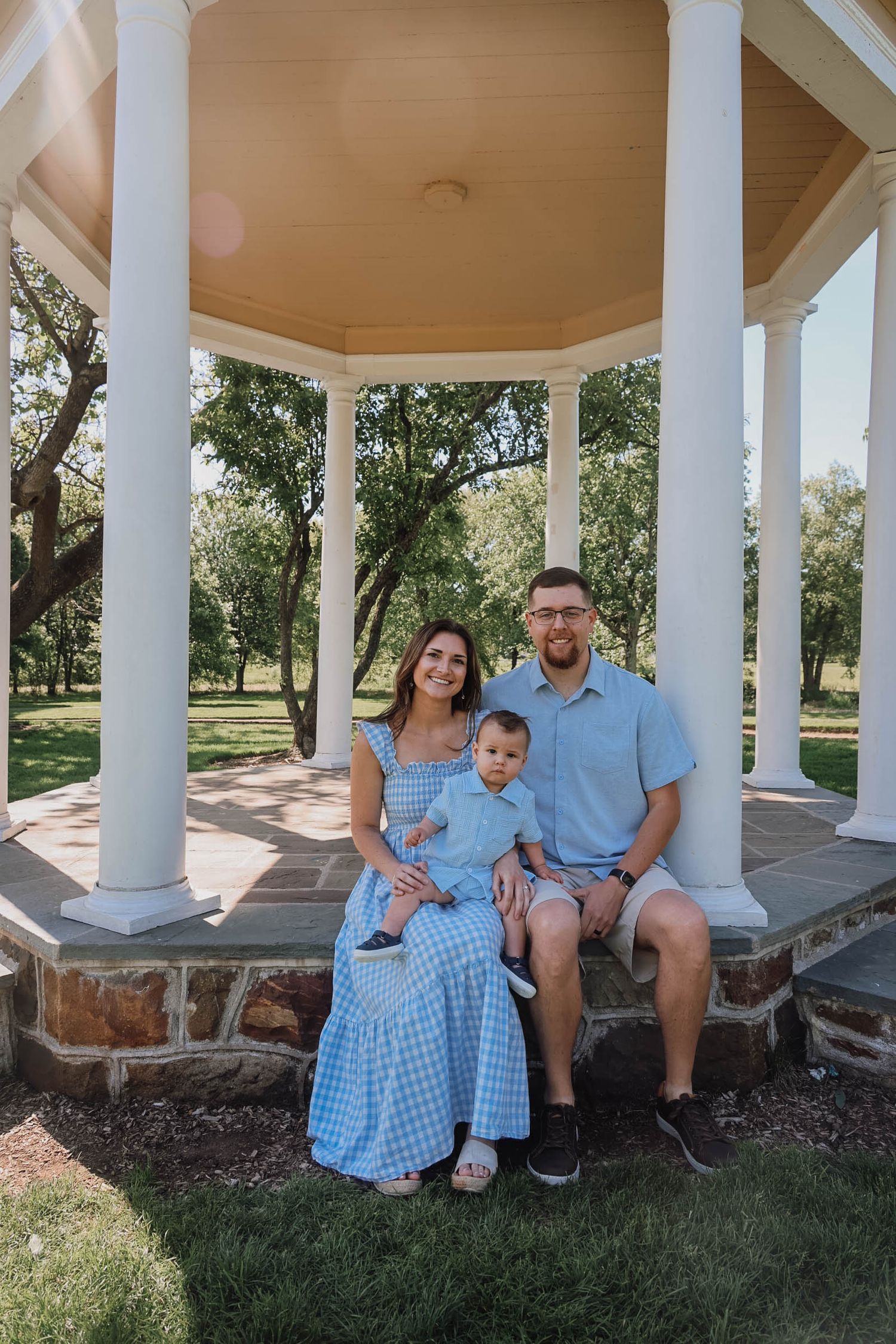 Family of three in blue outfits sitting under a white gazebo in a lush green park on a sunny day.