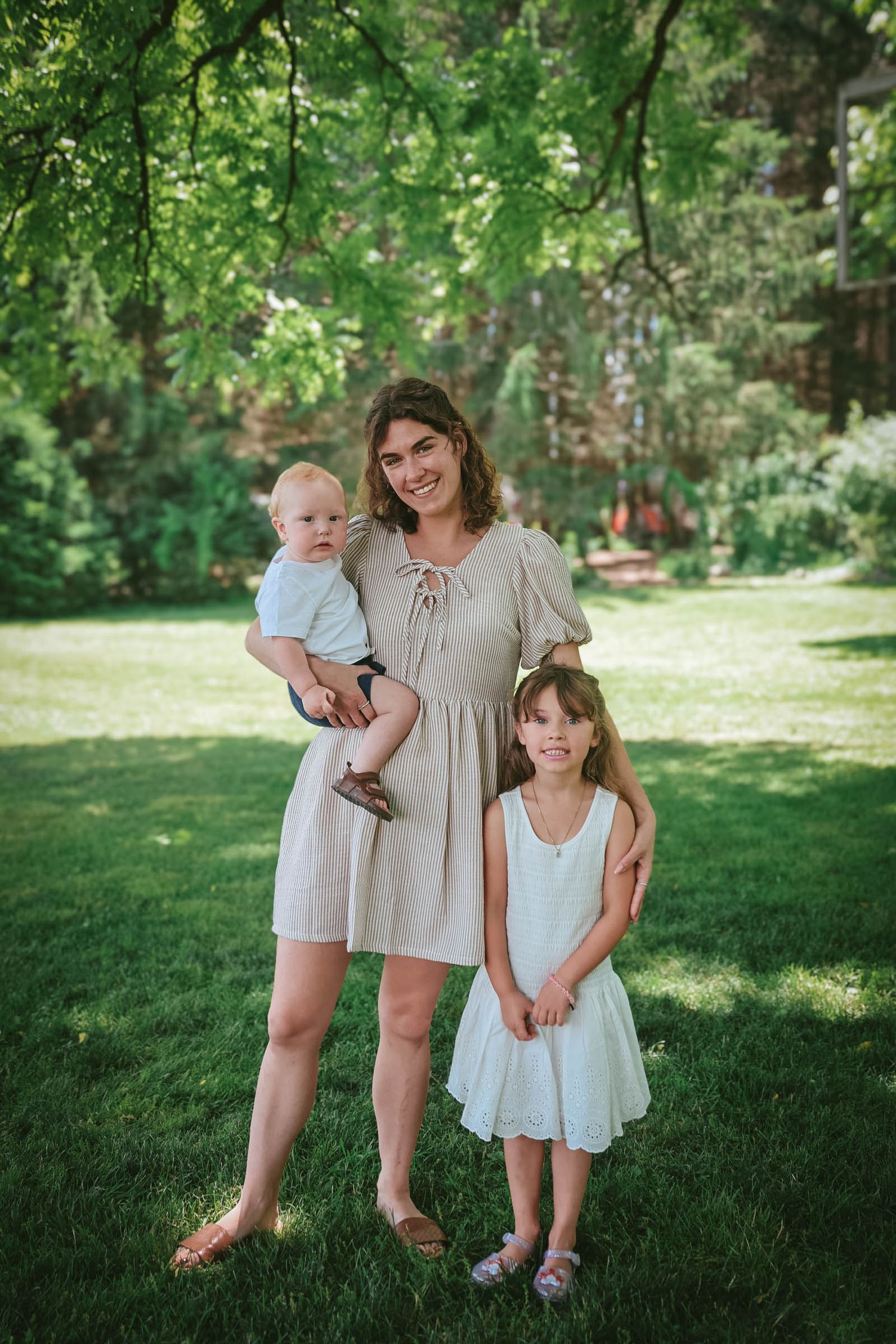 A smiling mother in a striped dress holds her baby while standing beside her young daughter in a lush green garden.