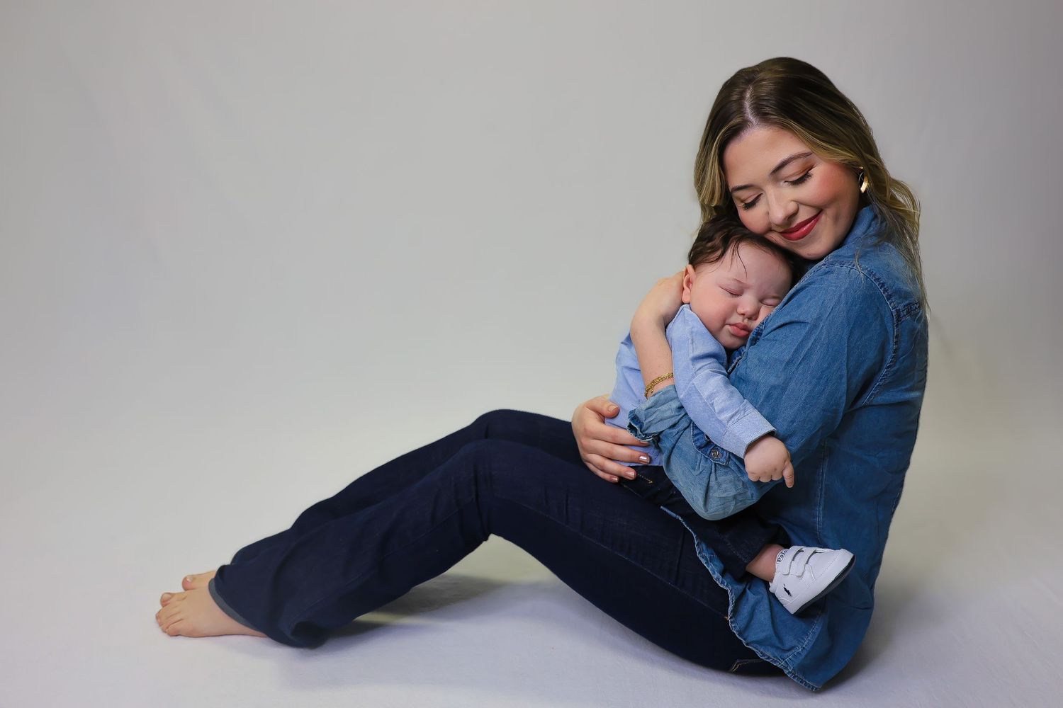 A smiling woman in denim sits on the floor cradling a sleeping newborn baby dressed in blue.
