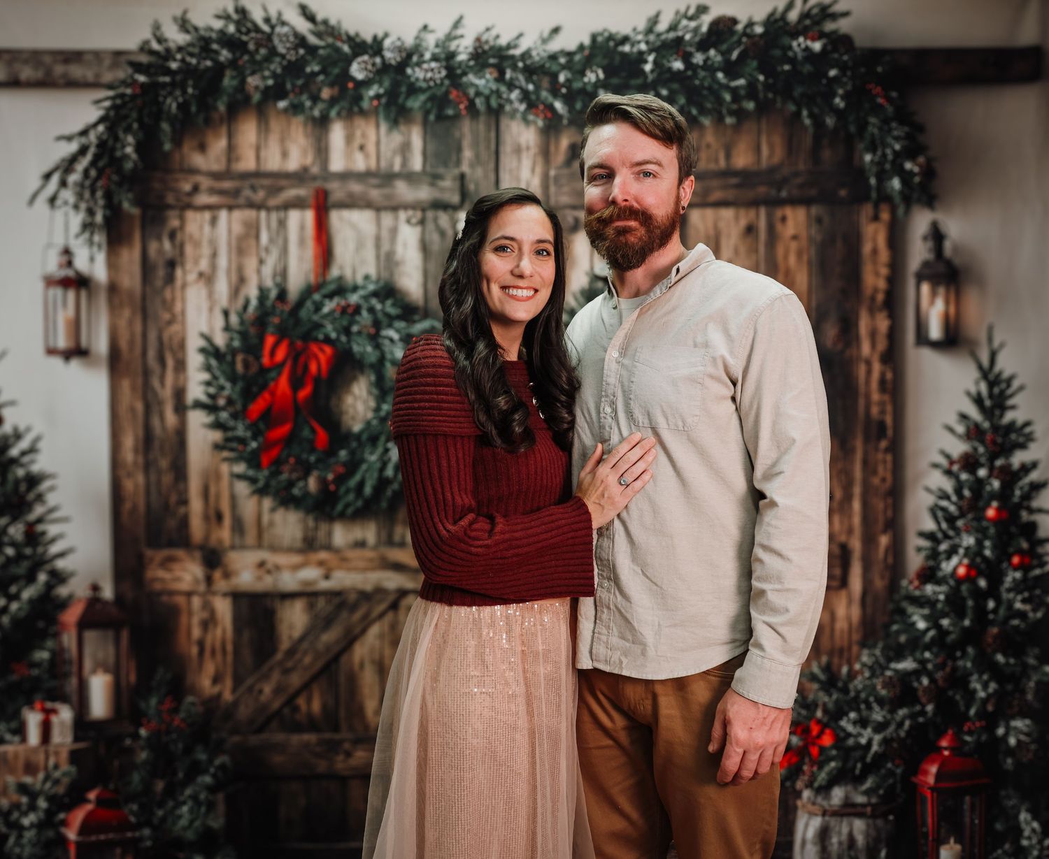 Couple posing together in front of rustic barn doors with Christmas wreaths and decorated trees in a festive holiday setting.