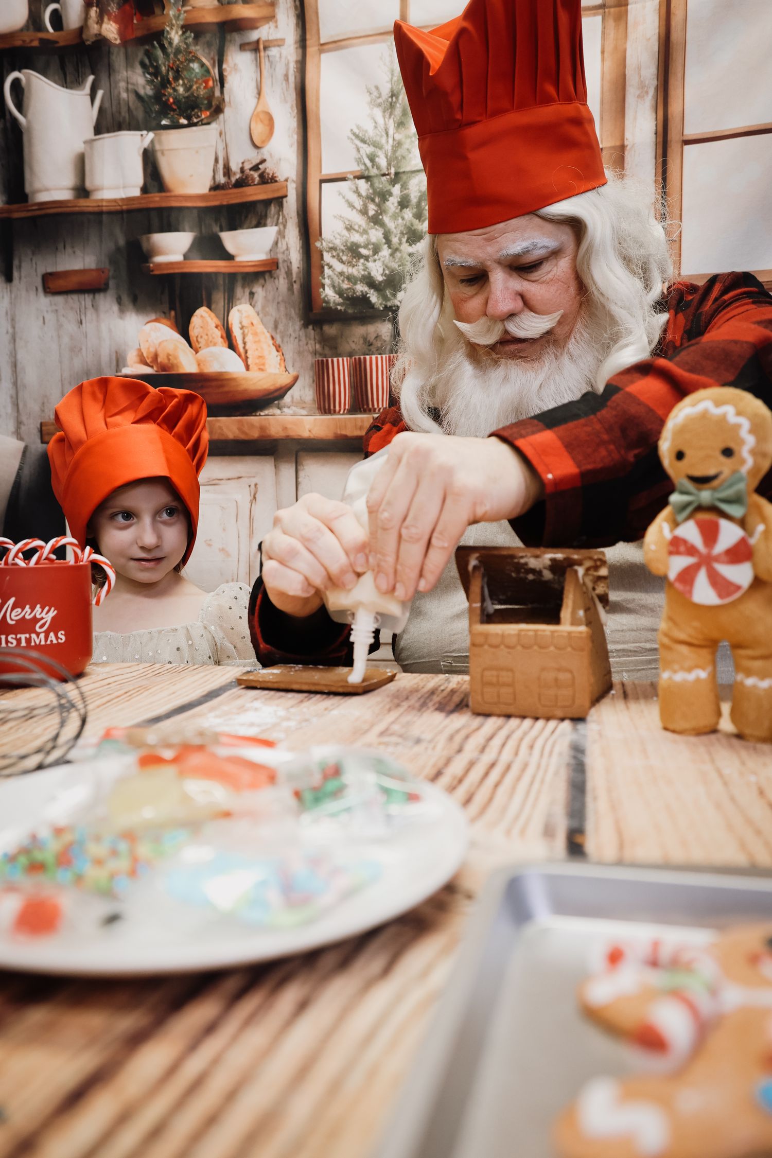 Santa Claus and child in red chef hats decorating a gingerbread house with icing at a Christmas kitchen table.