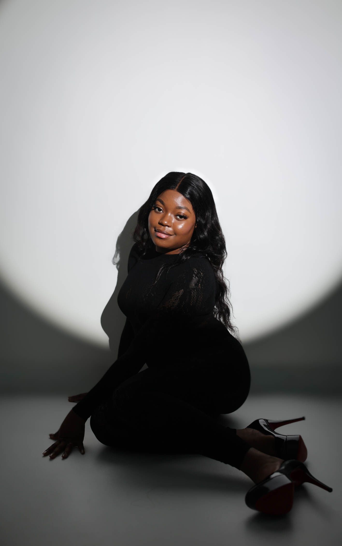 Woman in all-black outfit and heels sitting on floor, long wavy hair, posed against dramatic spotlight background.