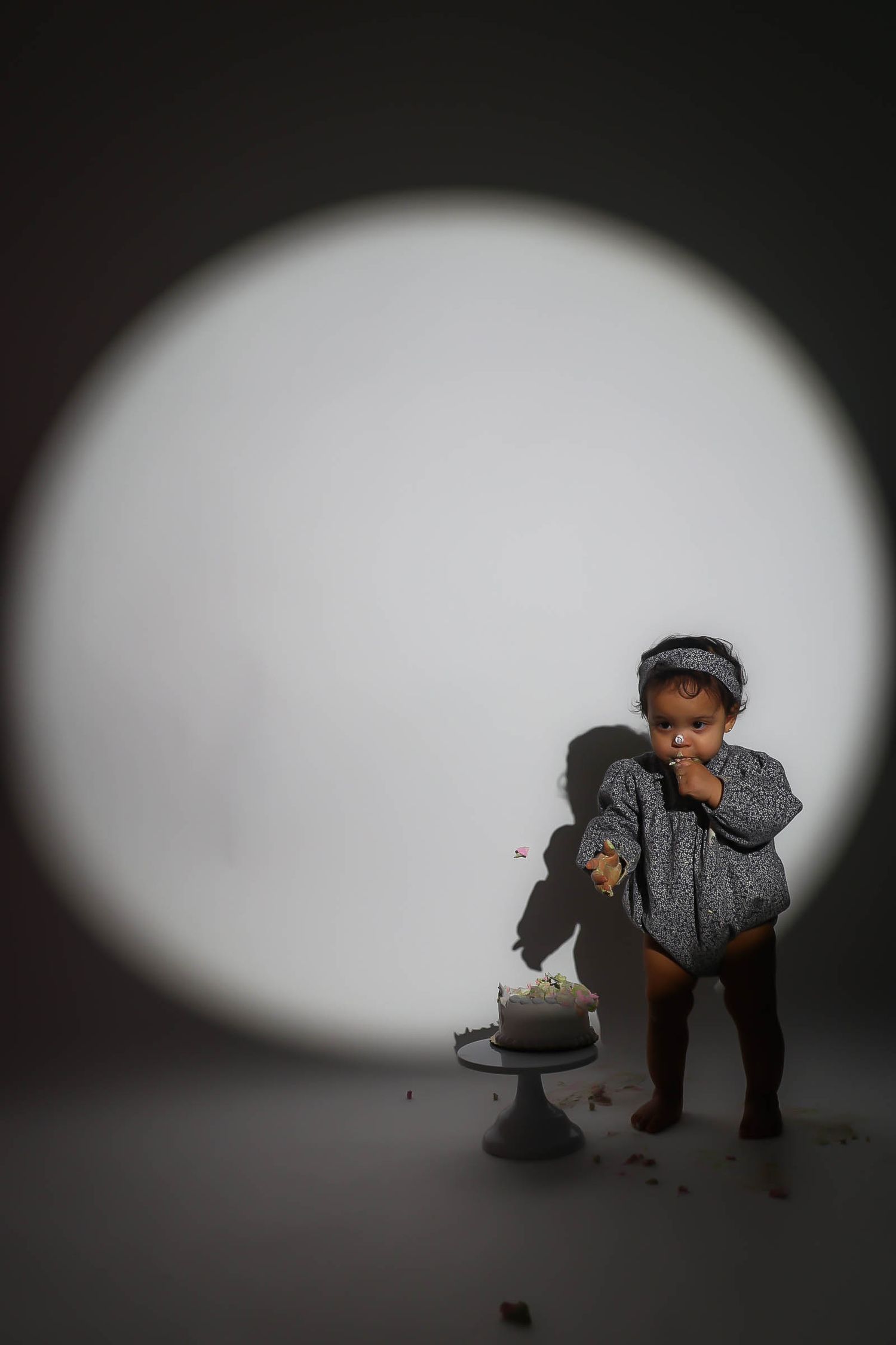 Cute toddler eating birthday cake wearing gray romper and headband in studio spotlight.