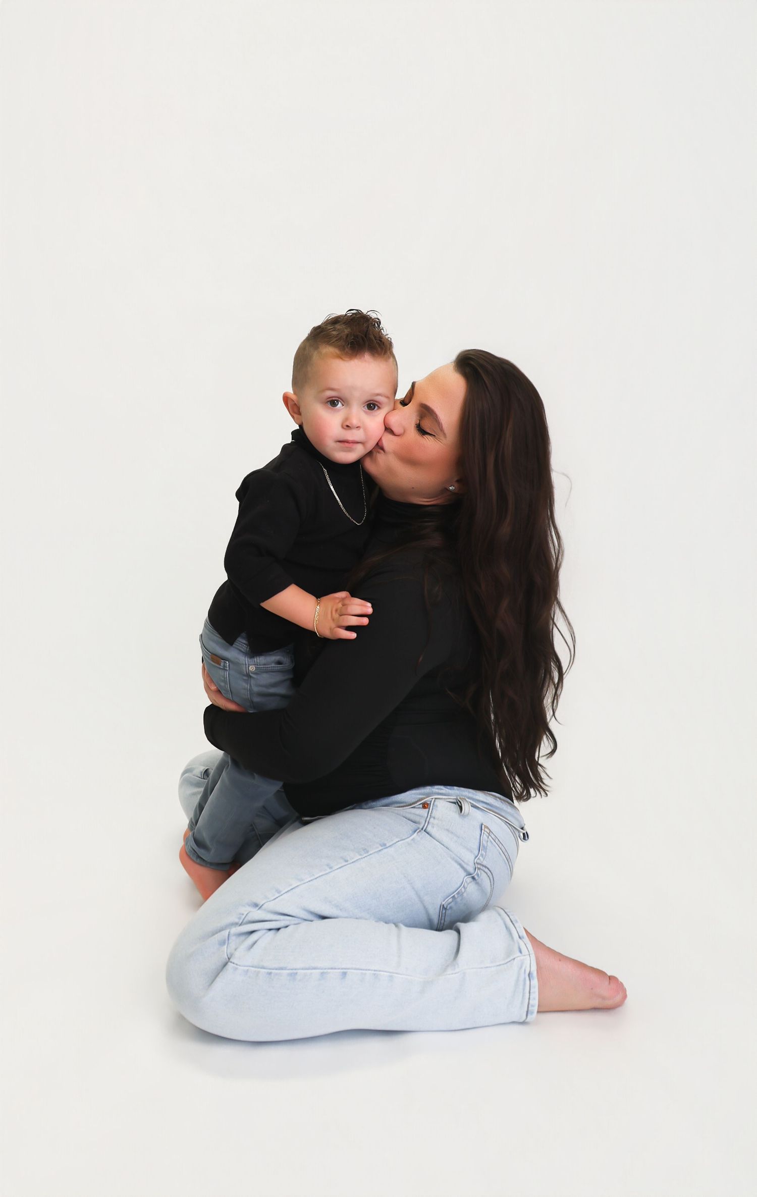 A woman with long dark hair sits on the floor kissing a toddler in a black hoodie, both wearing jeans.