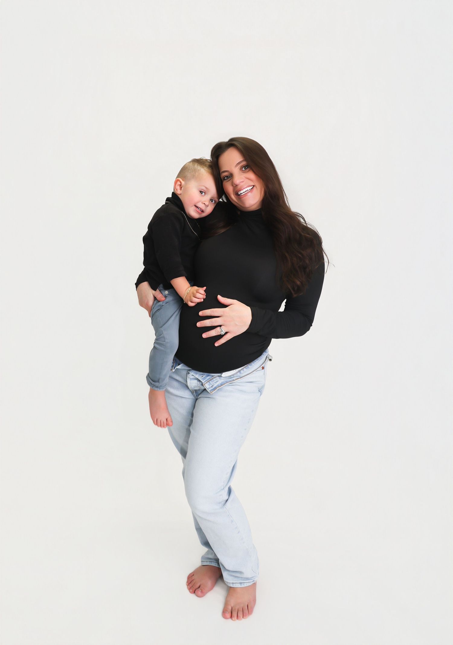 A smiling woman in black turtleneck and jeans holds a toddler in black against a white background.