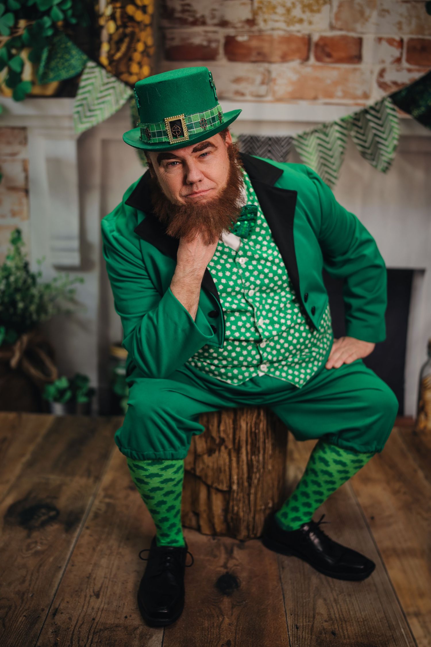 Man in green leprechaun costume with red beard sits on stump for St. Patrick's Day celebration.