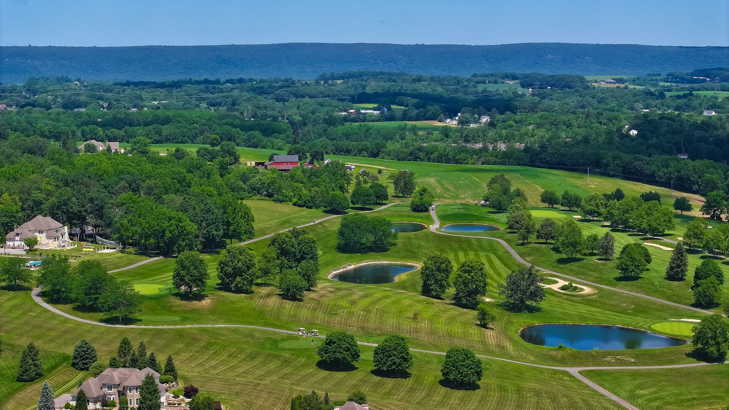 Aerial view of a lush green golf course with ponds, trees, and rolling hills under a clear blue sky.
