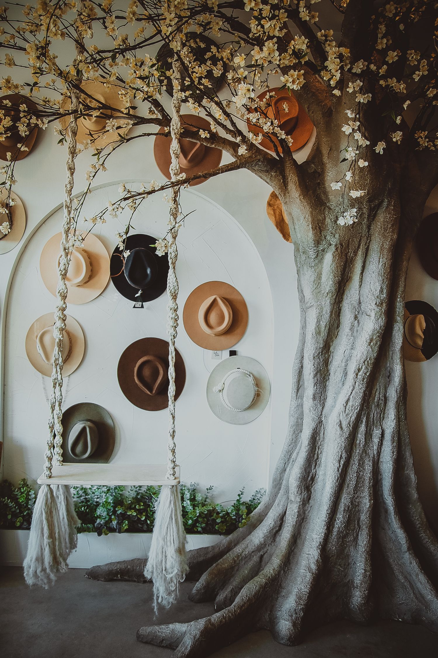 A decorative tree with white blossoms displaying various hats on a white wall beside a macrame swing.