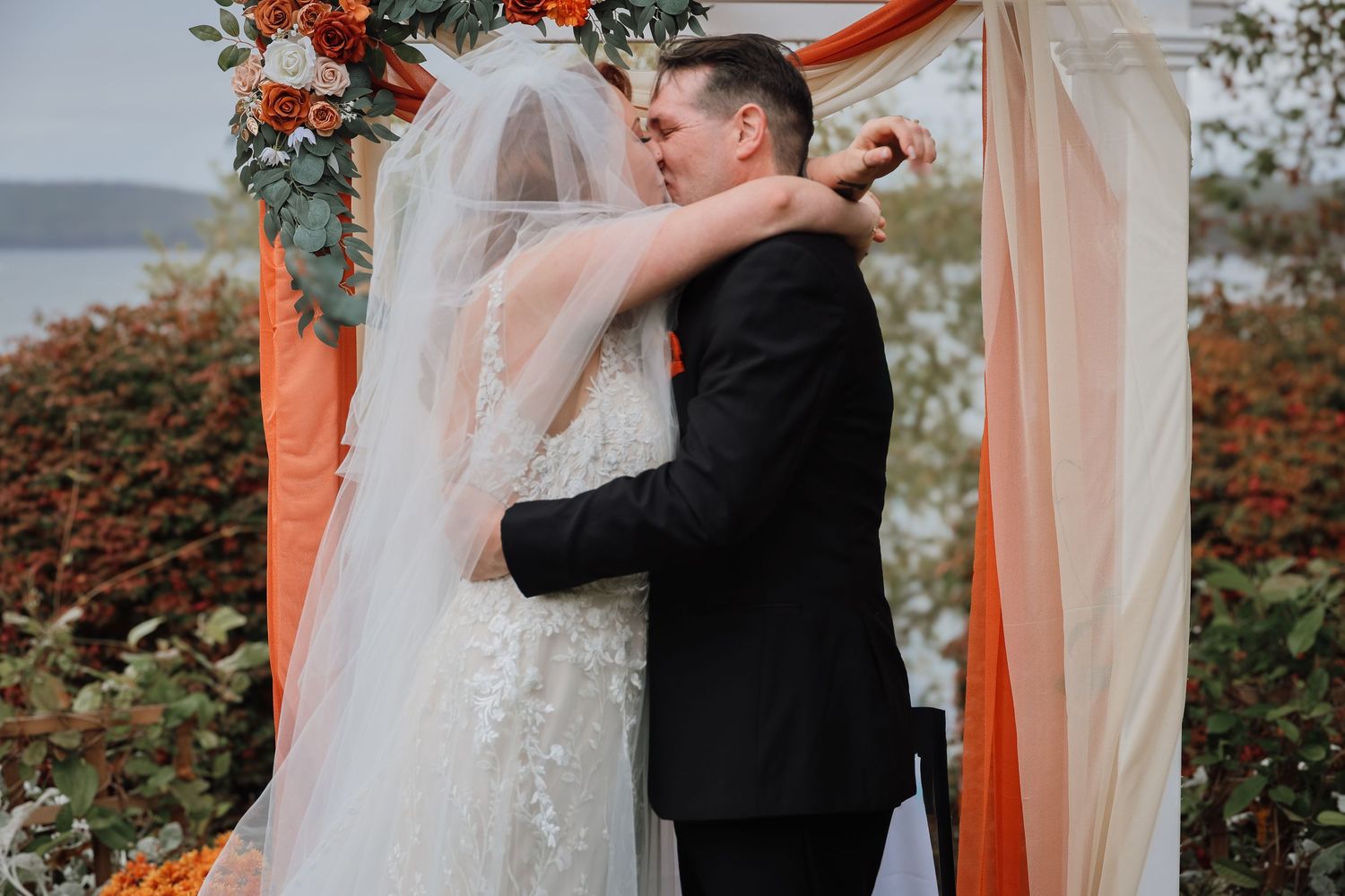 A romantic wedding ceremony under a coral-draped arch with floral decorations and greenery in an outdoor garden setting.
