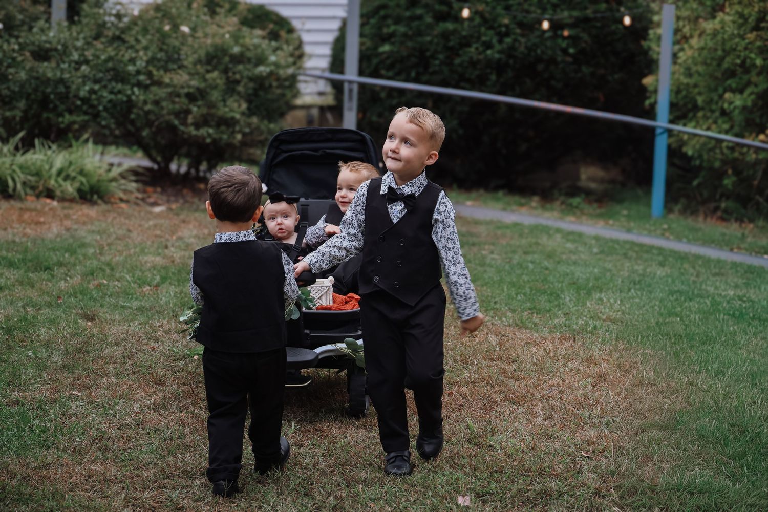 Two young children in formal attire play together near a black car in a backyard setting.