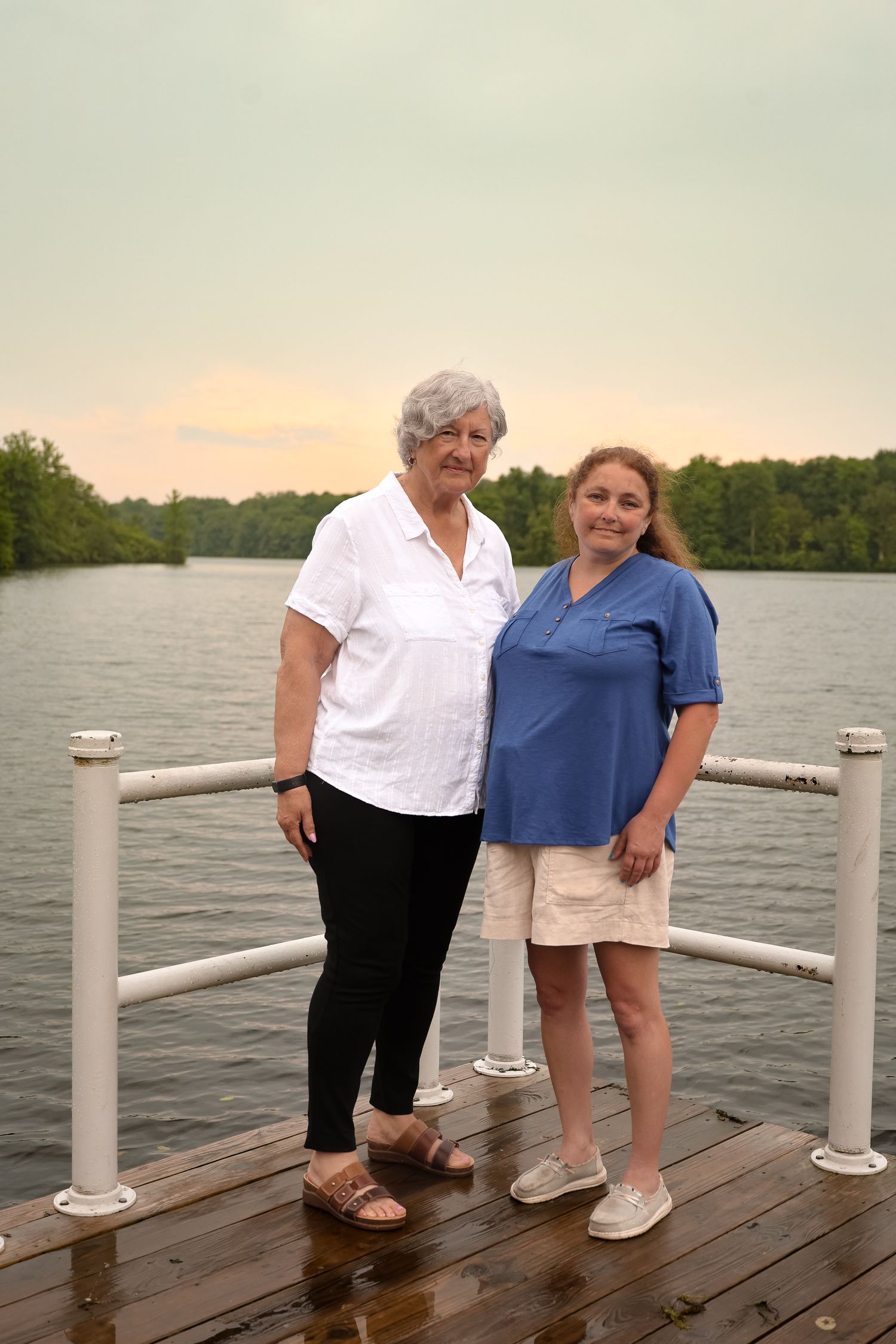 Two people stand together on a wooden dock by a serene lake at sunset, with trees visible in the background.