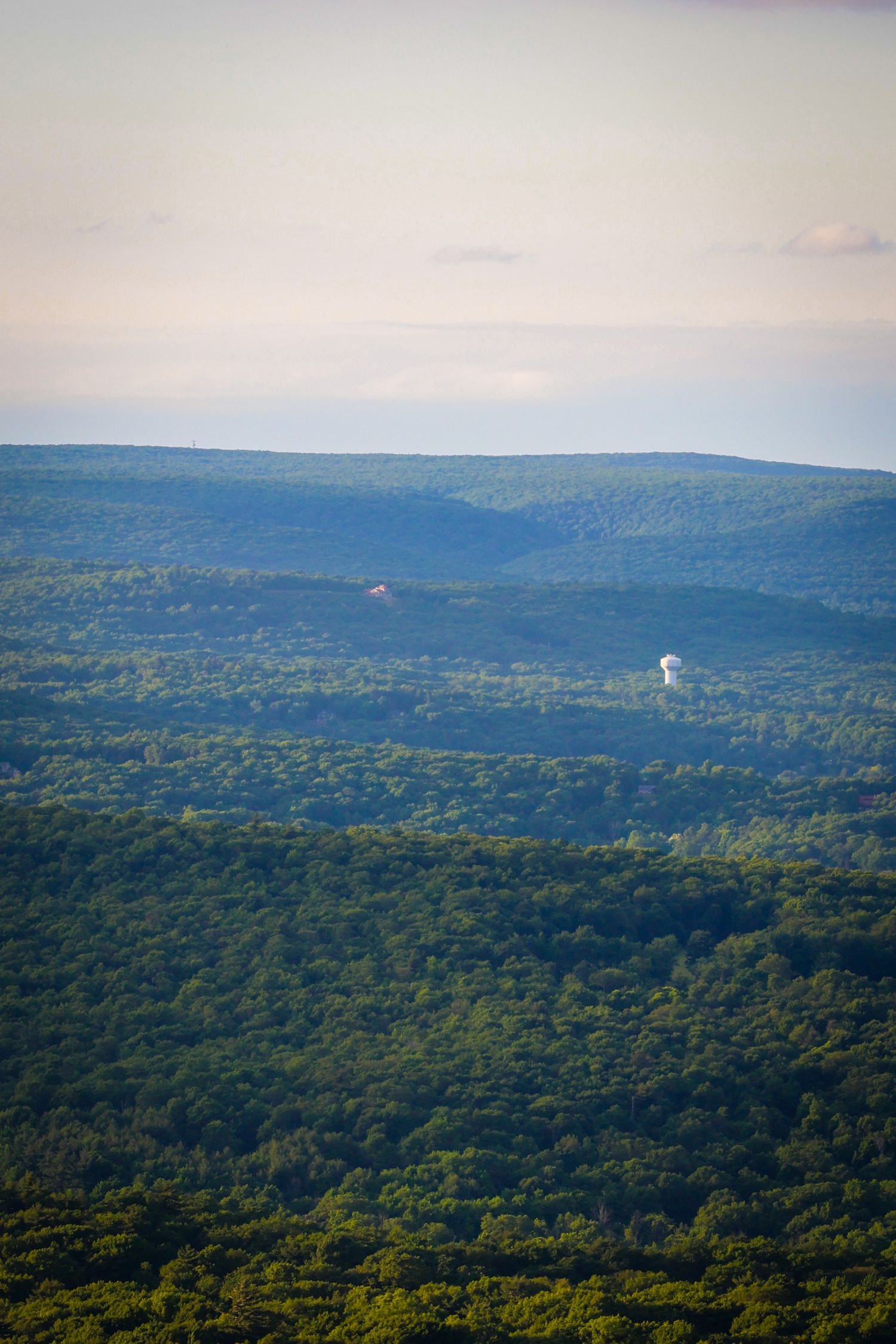 Rolling forested hills stretch to the horizon with a water tower visible in the distance against a hazy sky.