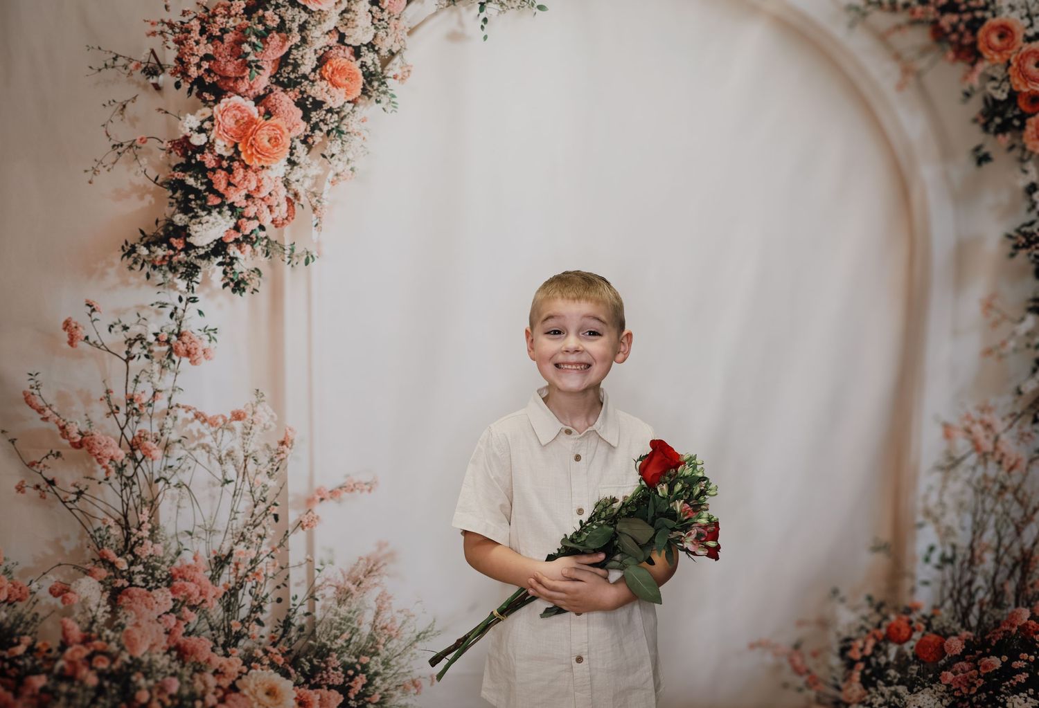 A child in white clothing holds a red rose under a floral arch decorated with peach-colored blooms.