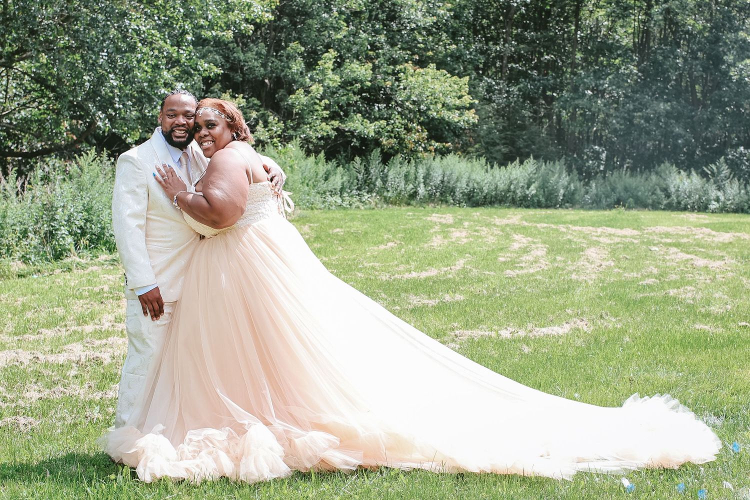 A couple in wedding attire embraces on a sunny day in a lush green outdoor garden setting.