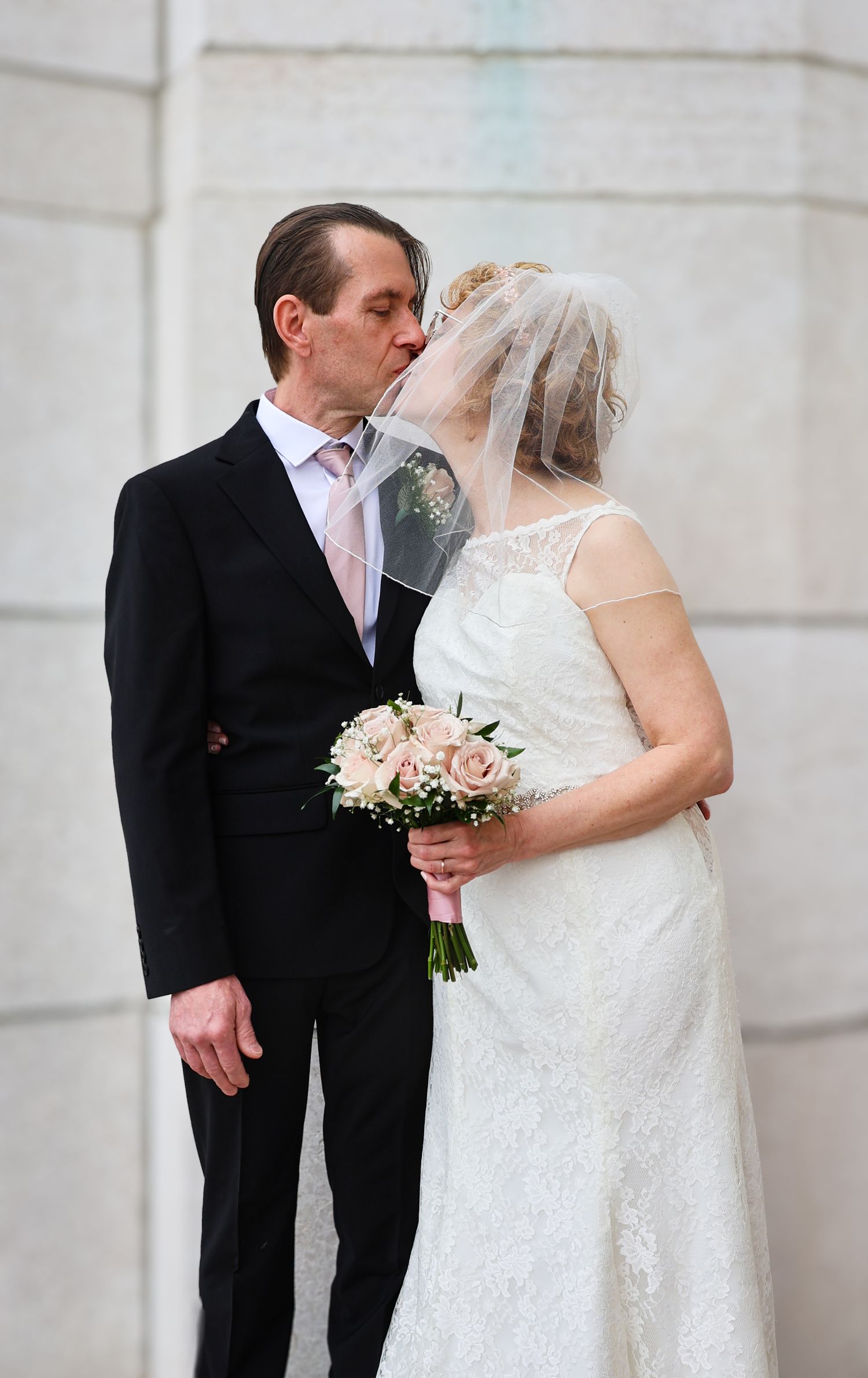 A romantic wedding moment captured as newlyweds share a kiss while bride holds a soft pink bouquet against white stone wall.