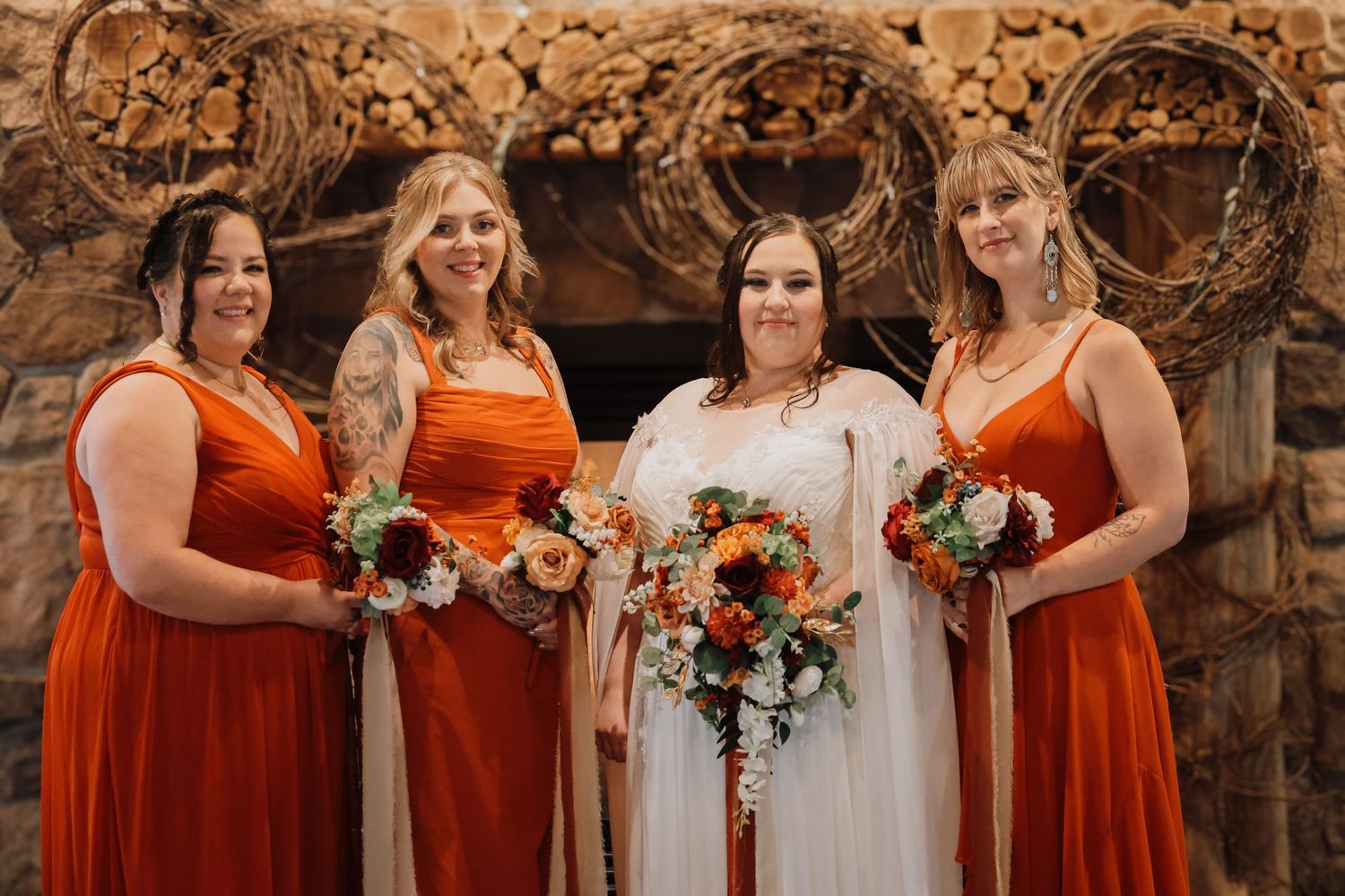 A wedding party poses together wearing orange and white dresses while holding floral bouquets against a stone wall.