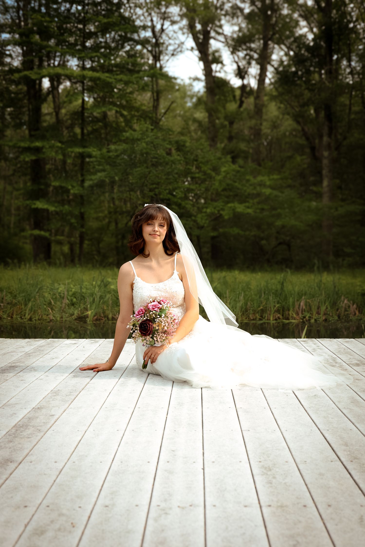 A bride in a white wedding gown sits gracefully on a wooden deck surrounded by lush green forest scenery.