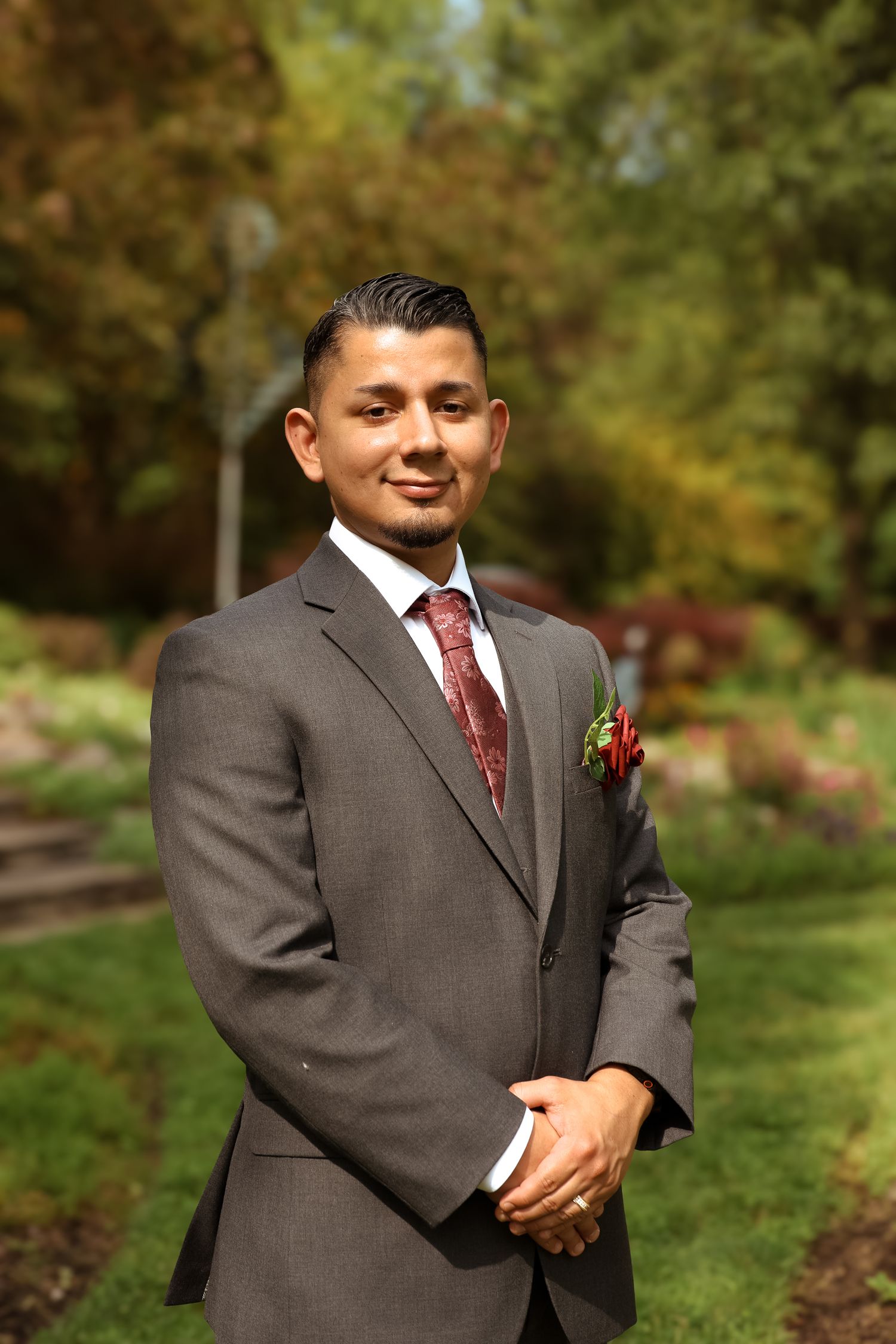 A groom in a brown suit with red tie and boutonniere poses outdoors in a garden setting during autumn.