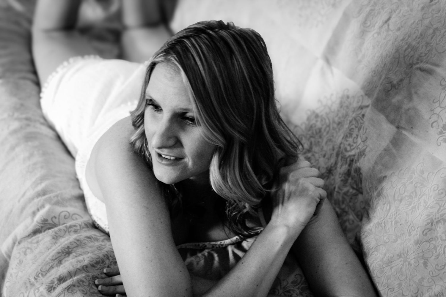 Black and white portrait of someone lying on bed with wavy hair and gentle smile looking to the side.