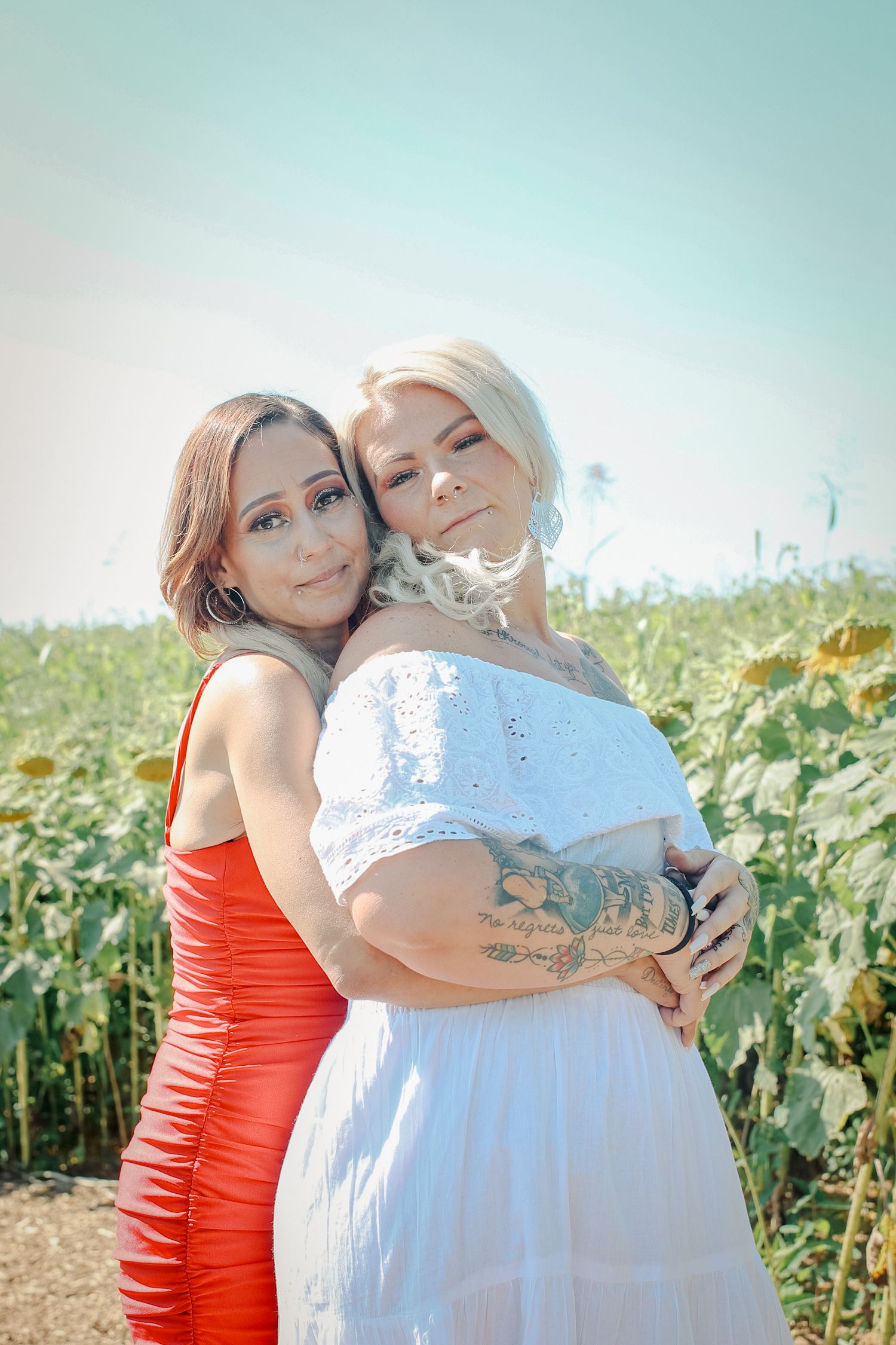 Two people in red and white outfits embrace in a sunflower field during a sunny outdoor photoshoot.