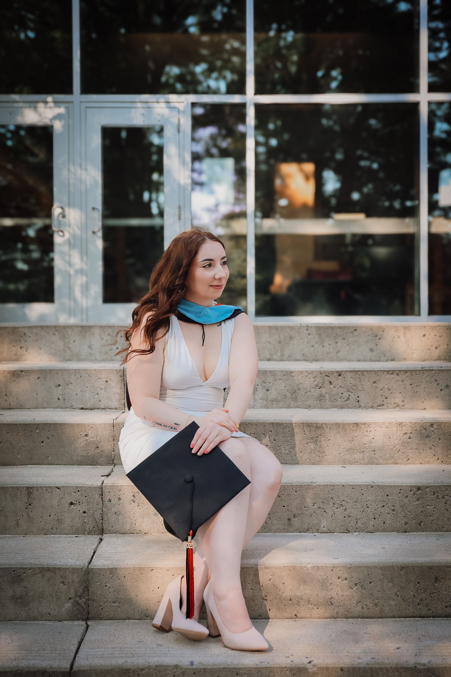 A graduate in a white dress sits thoughtfully on concrete steps near large windows on a sunny day.