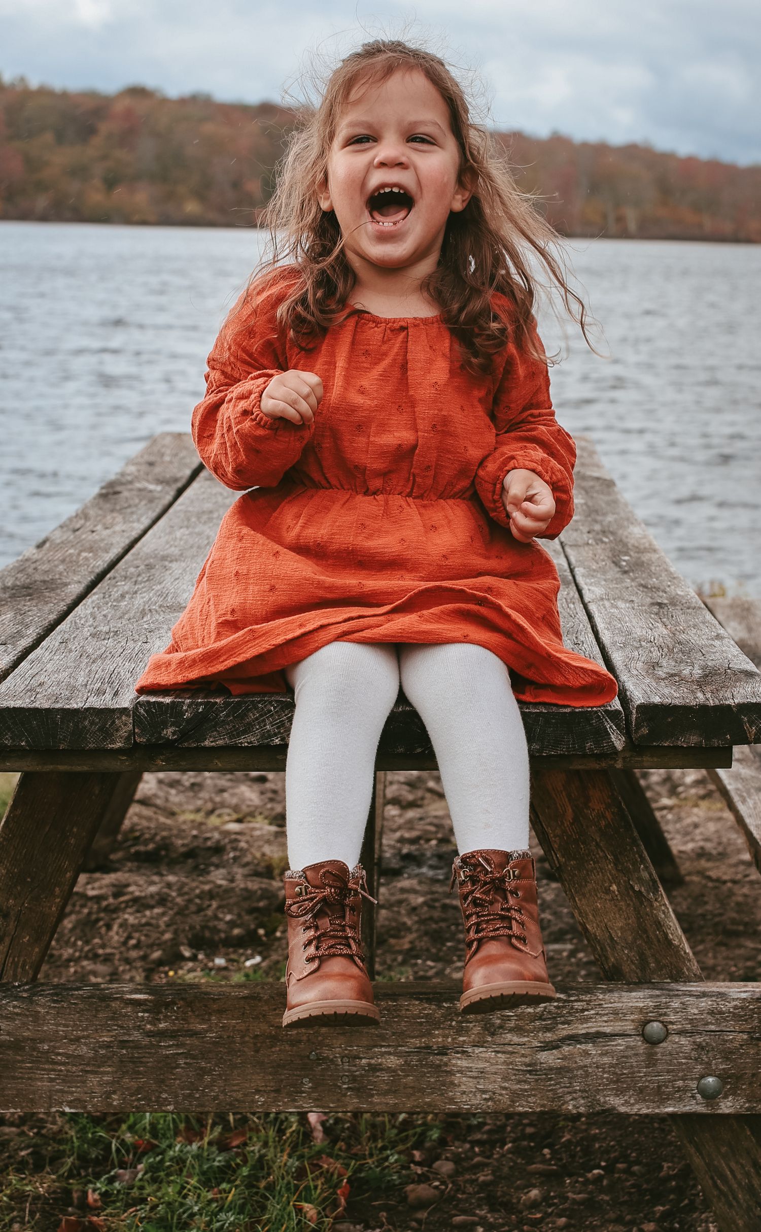 Child wearing orange sweater and brown boots sits on wooden dock by autumn lake, showing pure joy and laughter.