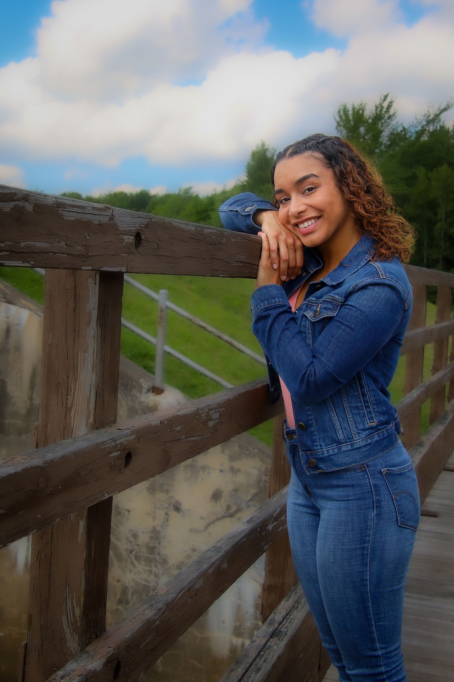 A person in denim poses casually against a wooden railing on a boardwalk with cloudy skies in the background.