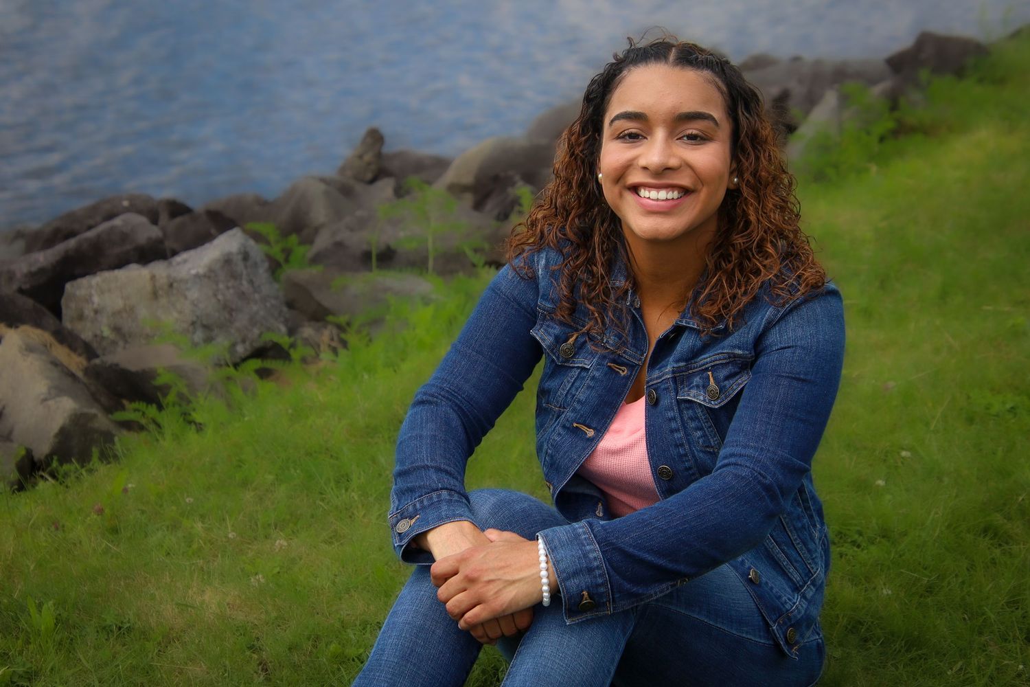A person in a denim jacket sits outdoors on grassy terrain with a scenic mountain landscape in the background.