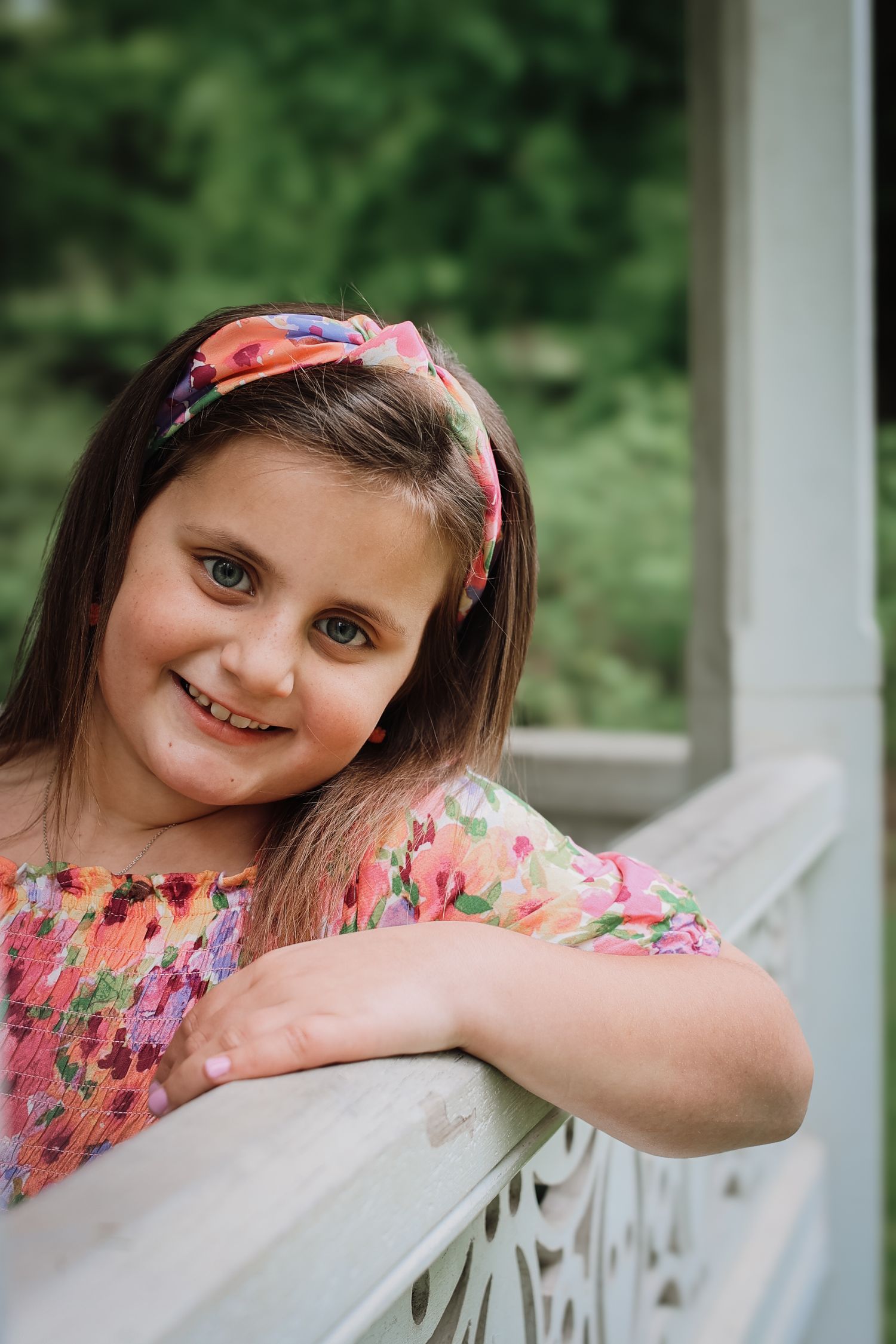 A child in a floral dress and pink headband leans on a white railing while smiling outdoors.