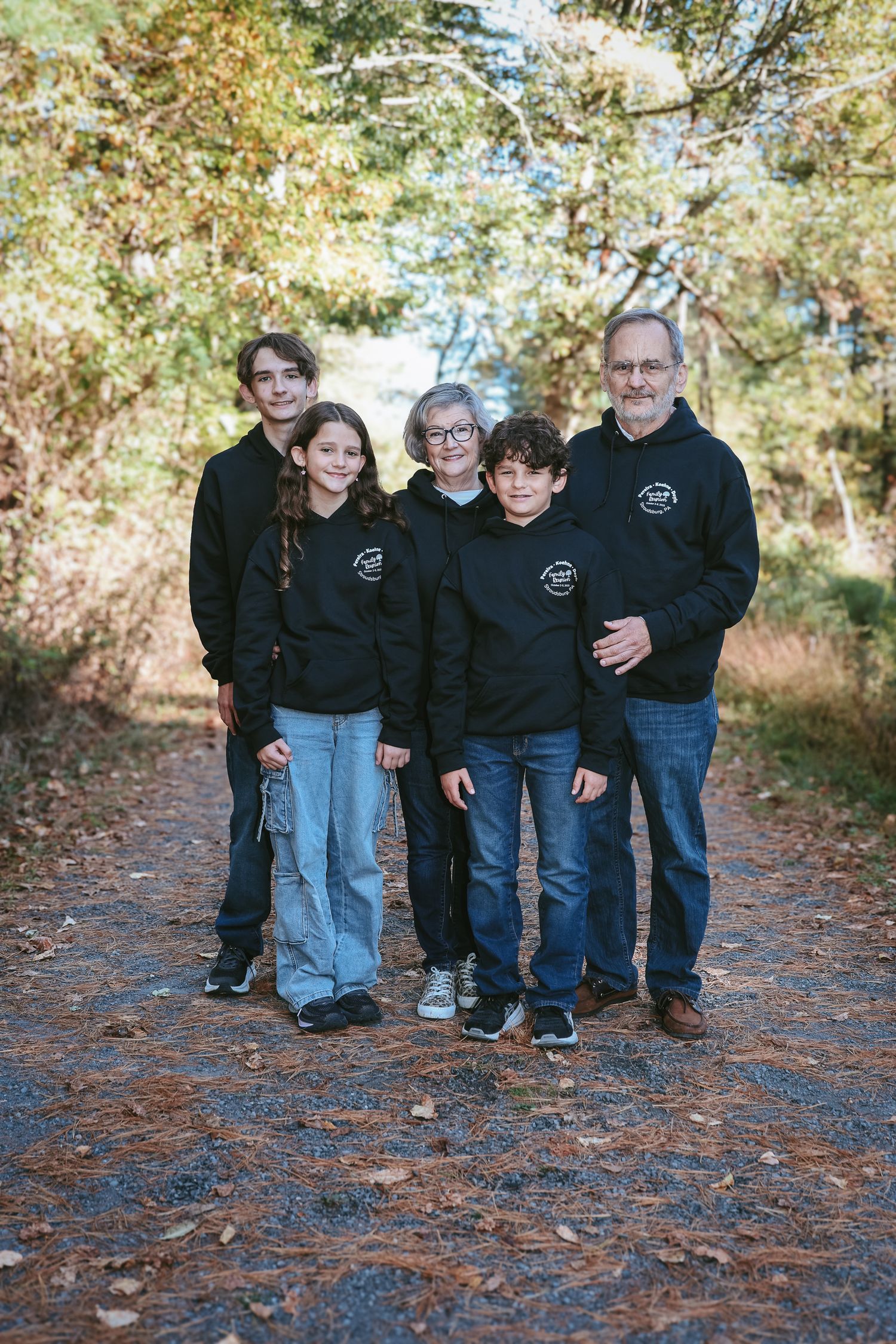 A group wearing matching black sweatshirts poses together on a leaf-covered forest path in autumn.