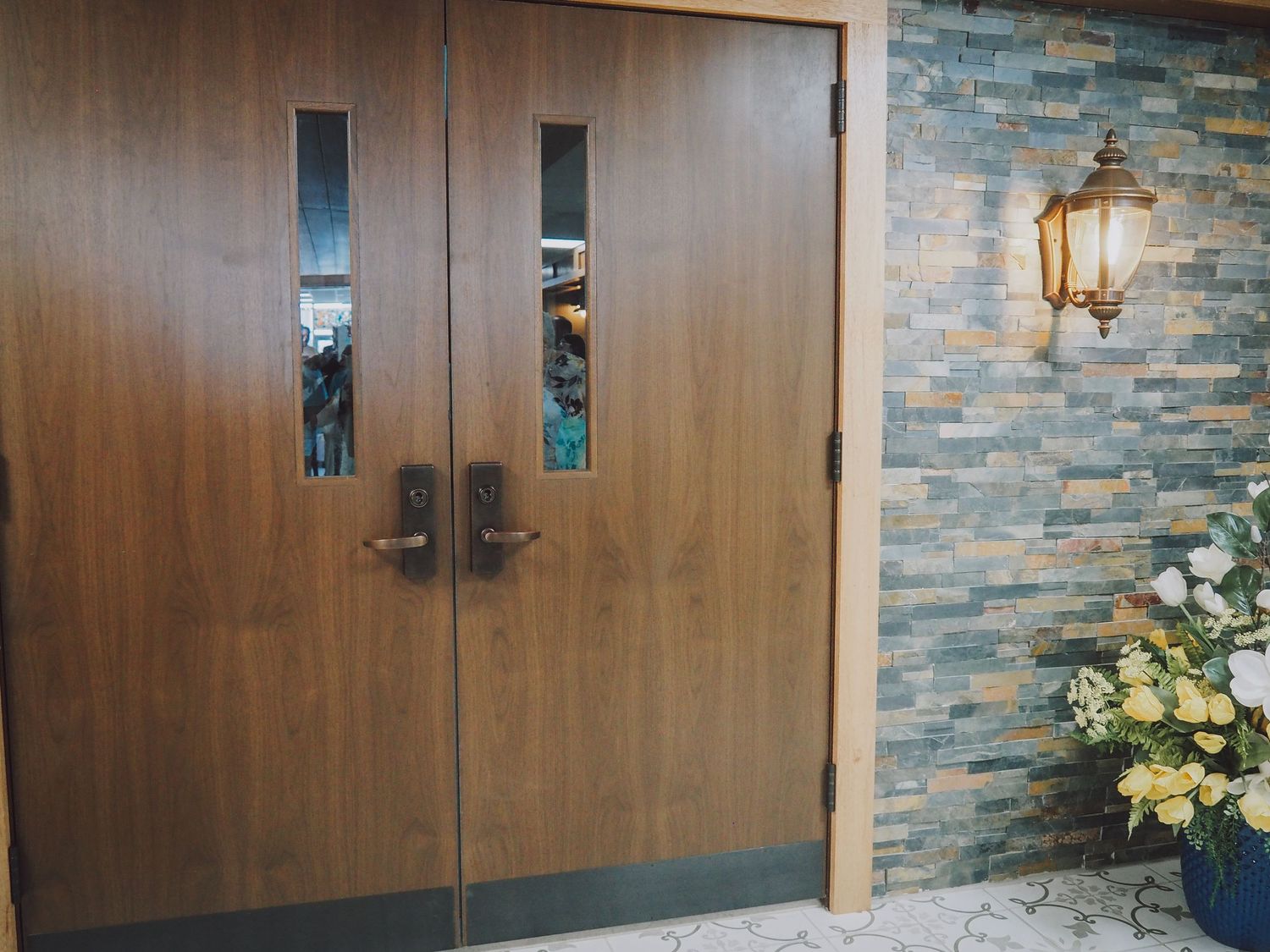 Double wooden doors with vertical windows and black handles against stone wall with decorative lantern and floral arrangement.