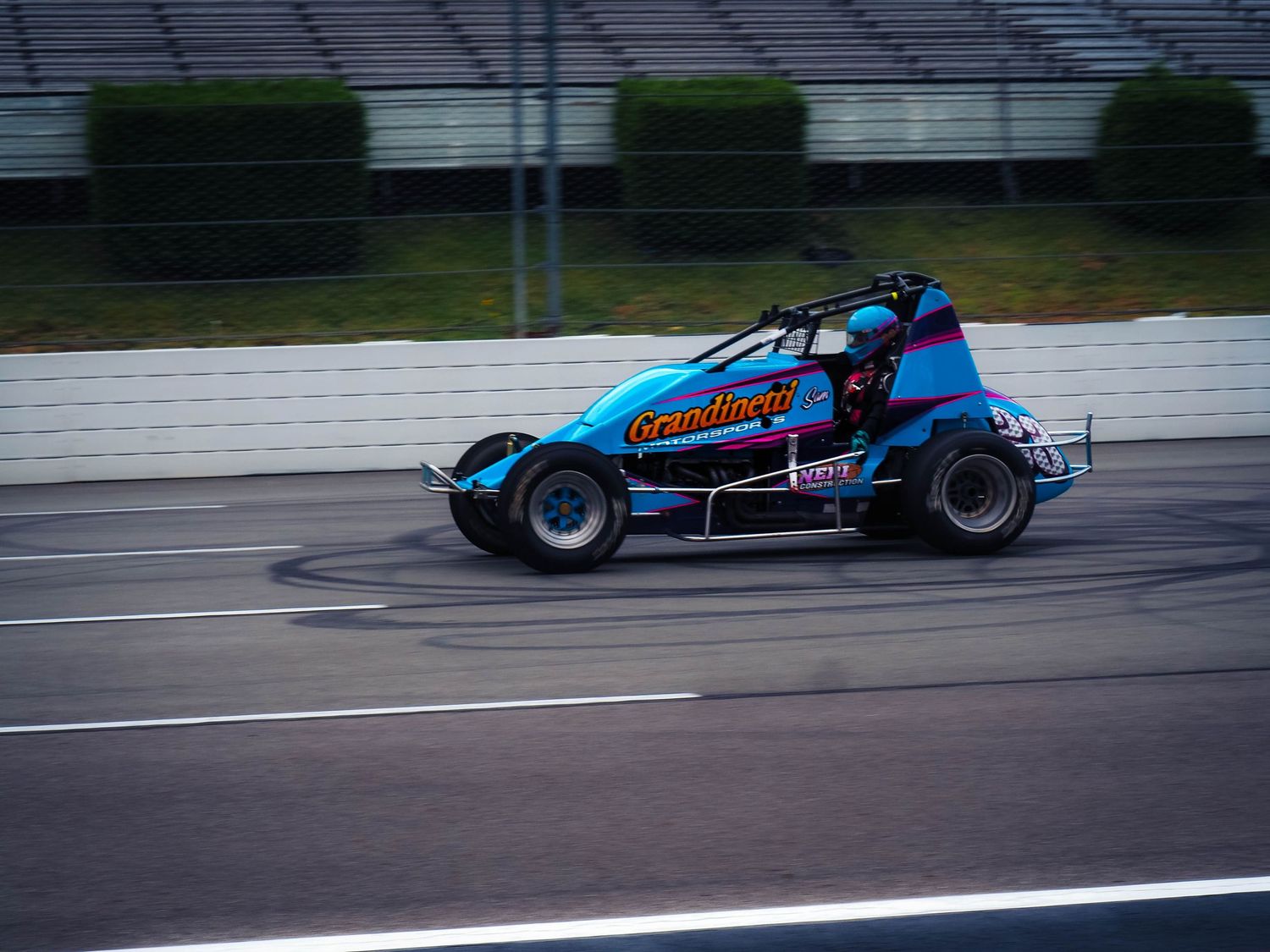 Blue race car speeds along a wet racetrack during a motorsport competition.