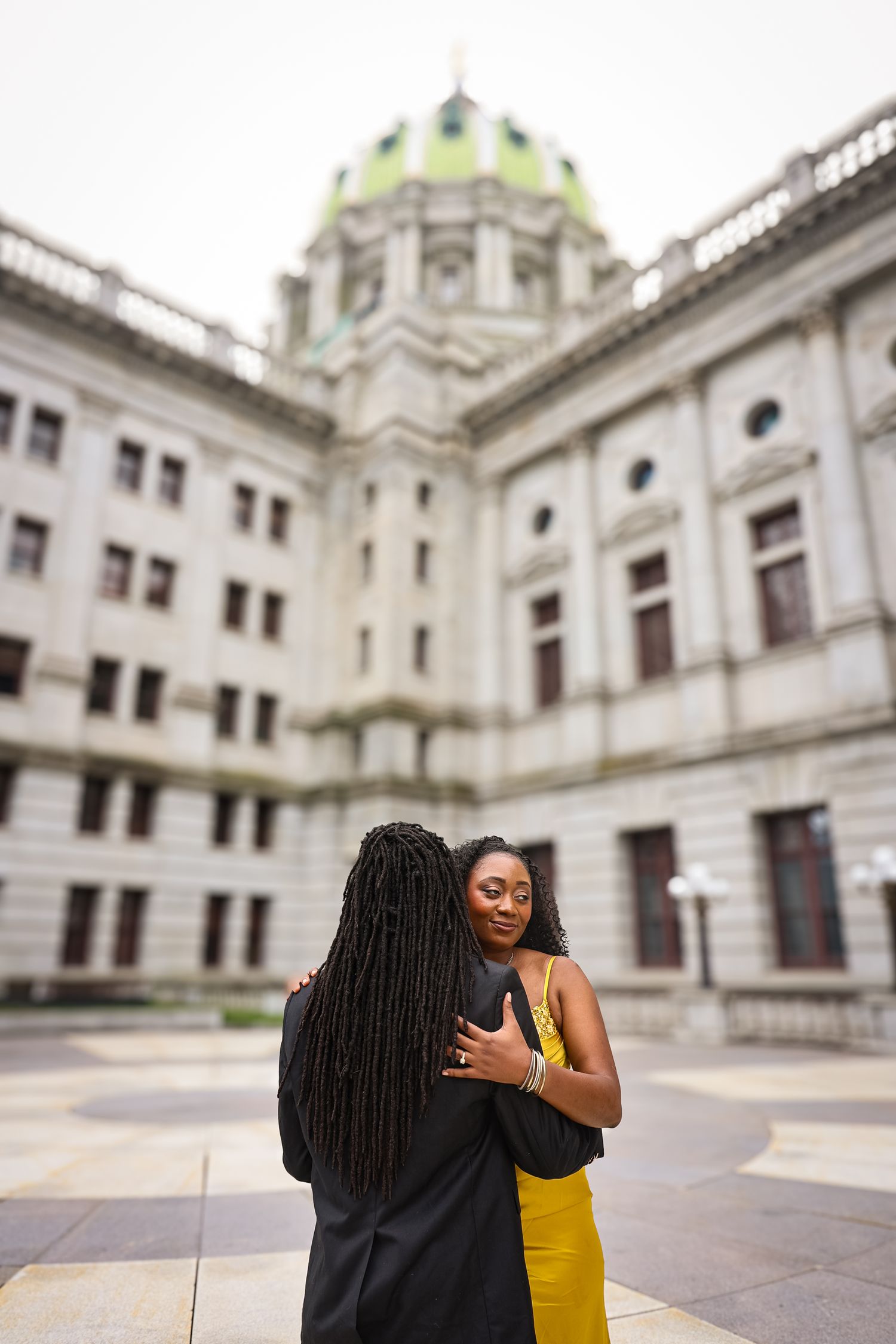 Two people share an embrace in front of a grand white historic building with ornate architecture.
