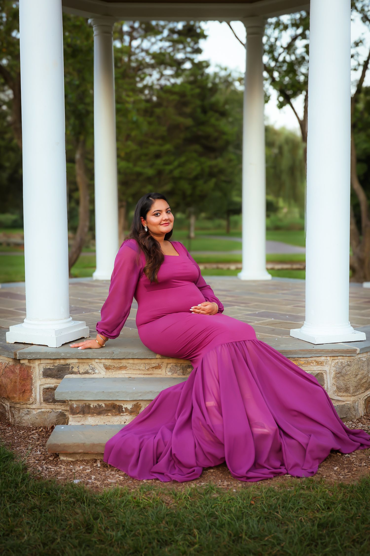 A woman in a flowing magenta gown poses elegantly on stone steps surrounded by white columns in an outdoor setting.