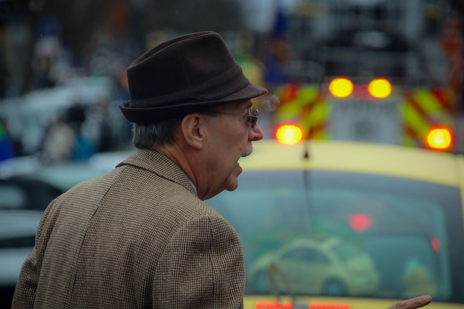 A person wearing a brown coat and hat walks past a row of parked cars on a city street at dusk.