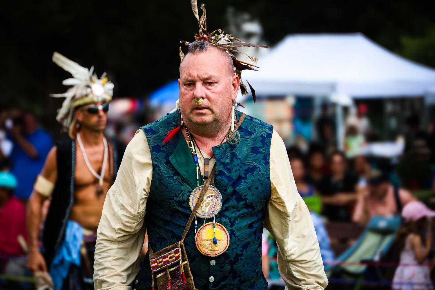 A performer in medieval Renaissance faire costume with a feathered hat and pendant necklace interacts with festival attendees.