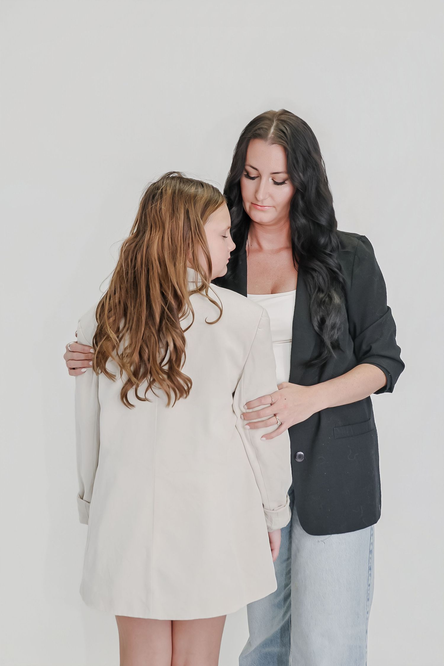 A woman in a black blazer tenderly embraces a young girl with long wavy hair in a white blazer against a light background.